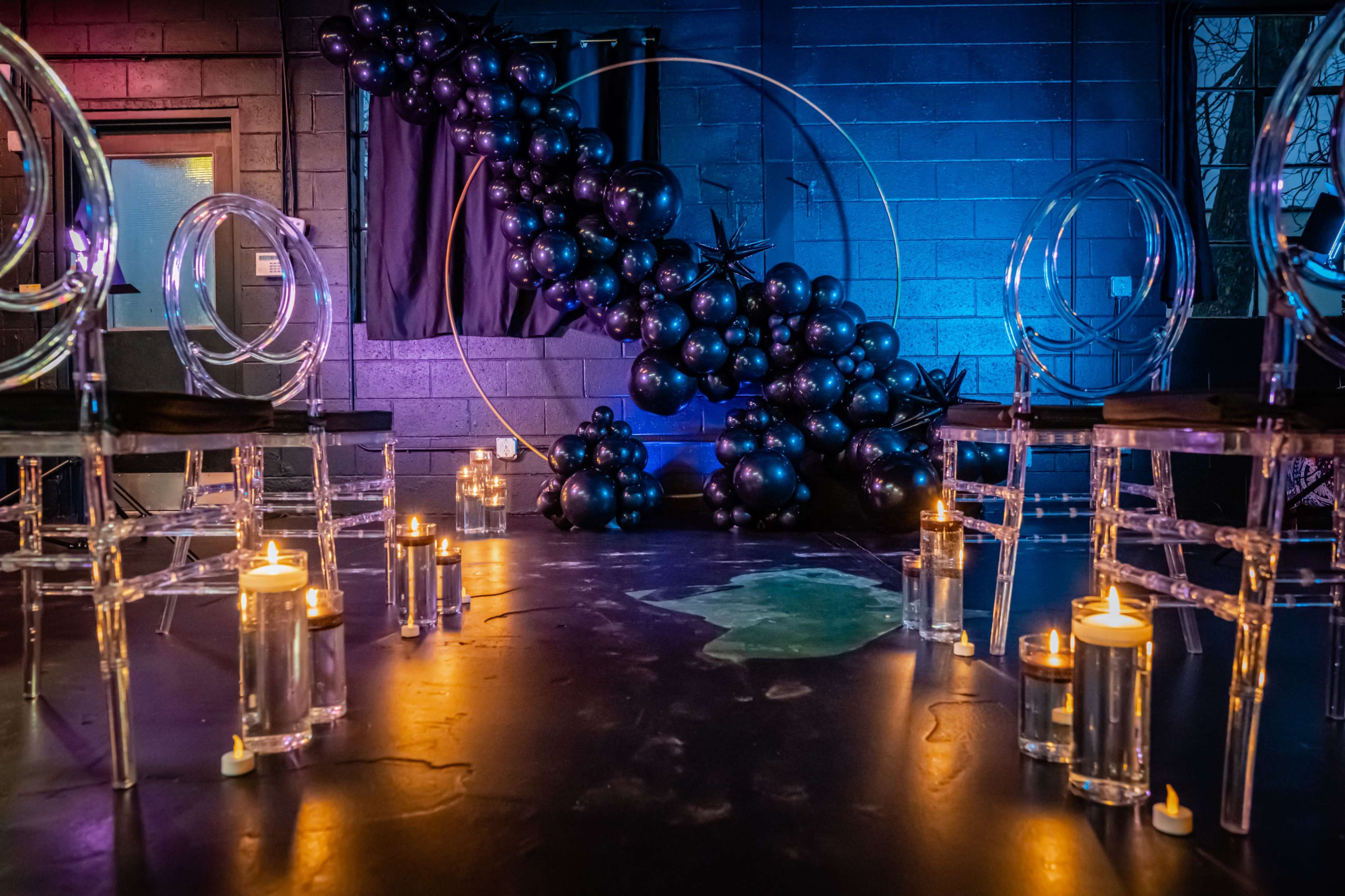 The scene features a darkened room with clear chairs arranged in a circle, surrounded by glowing candles and an artistic arrangement of black balloons.