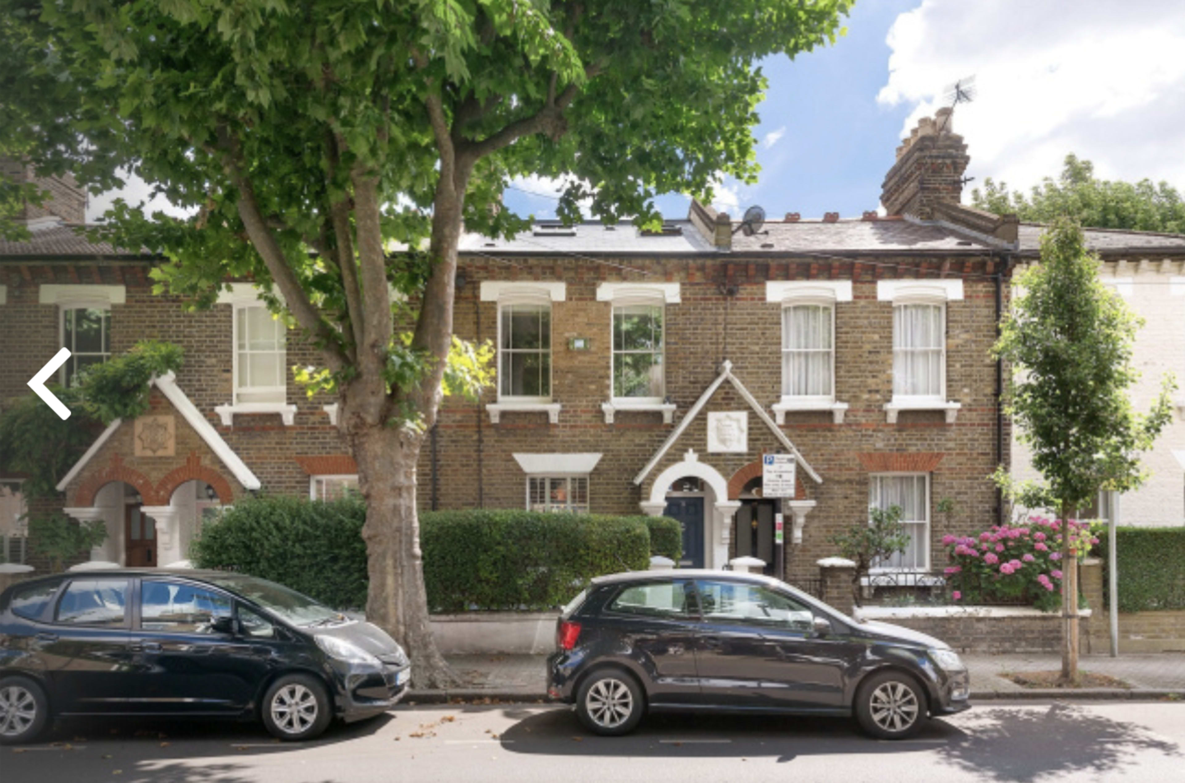 The image shows a row of brick terraced houses on a tree-lined street, with parked cars along the curb and flowering bushes in front of the homes.