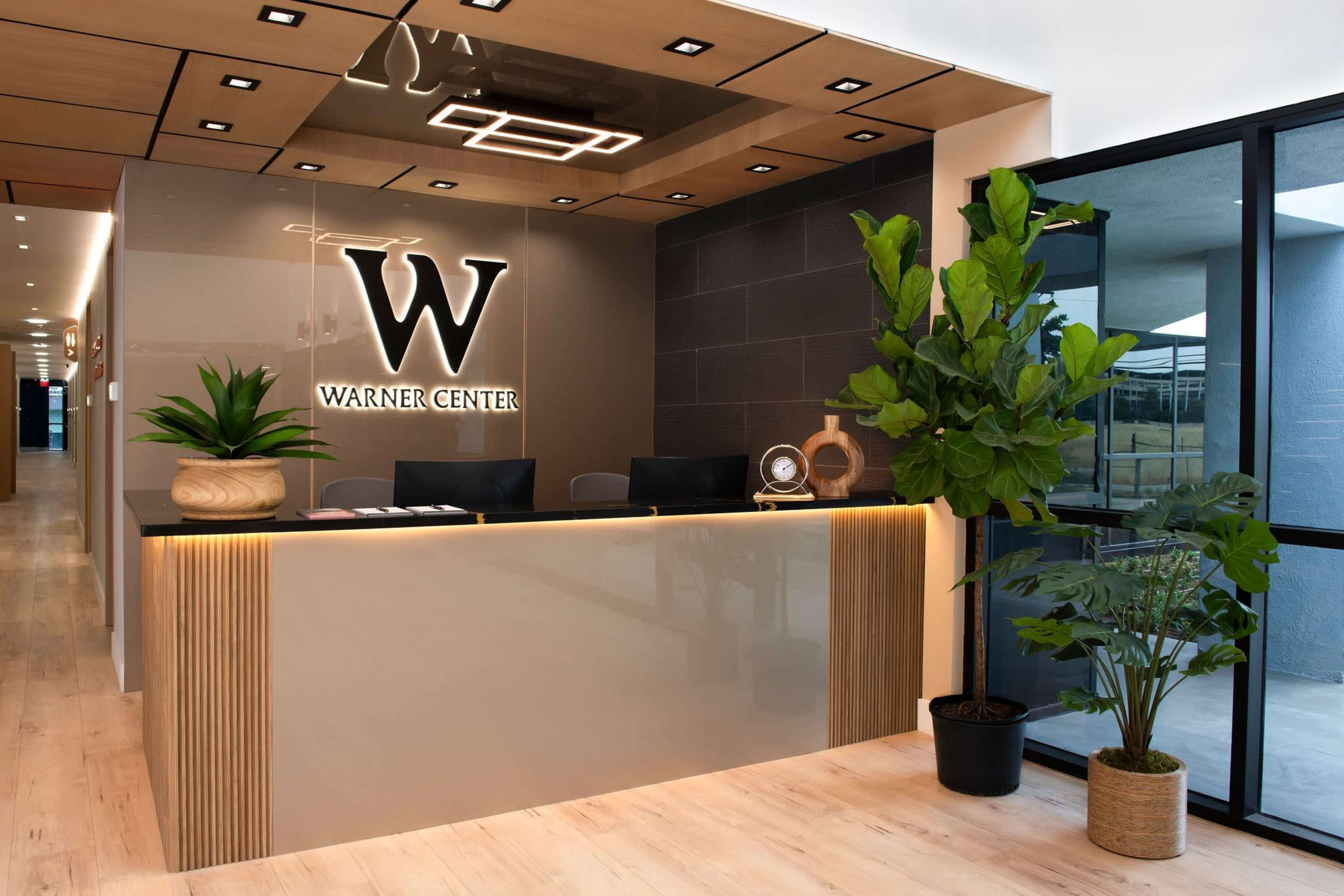 The image shows a modern reception area featuring a sleek front desk with the "Warner Center" logo, surrounded by decorative plants and large windows.