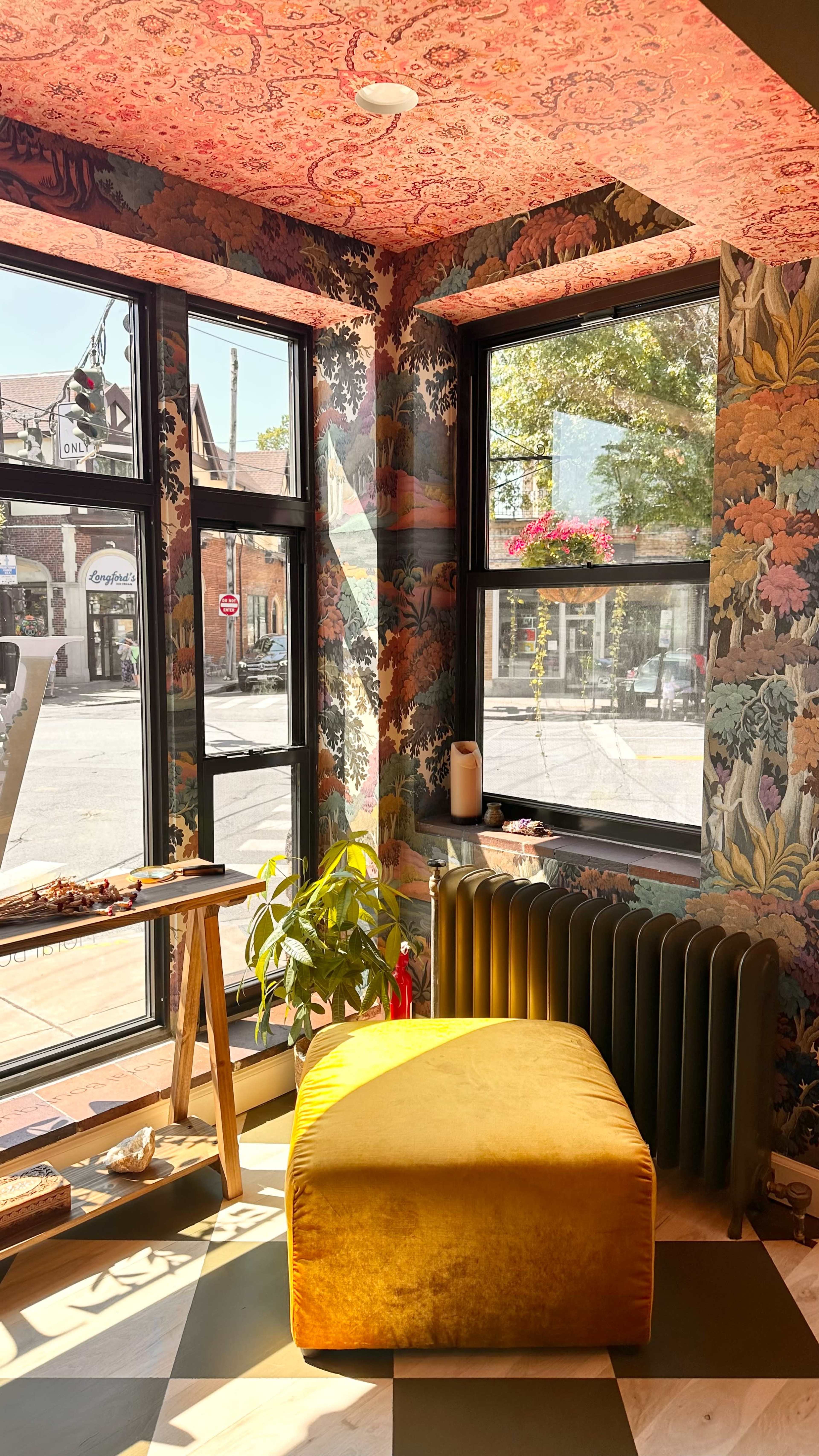 A cozy corner of a room with vibrant floral wallpaper, a yellow ottoman, a wooden table, and large windows letting in natural light.
