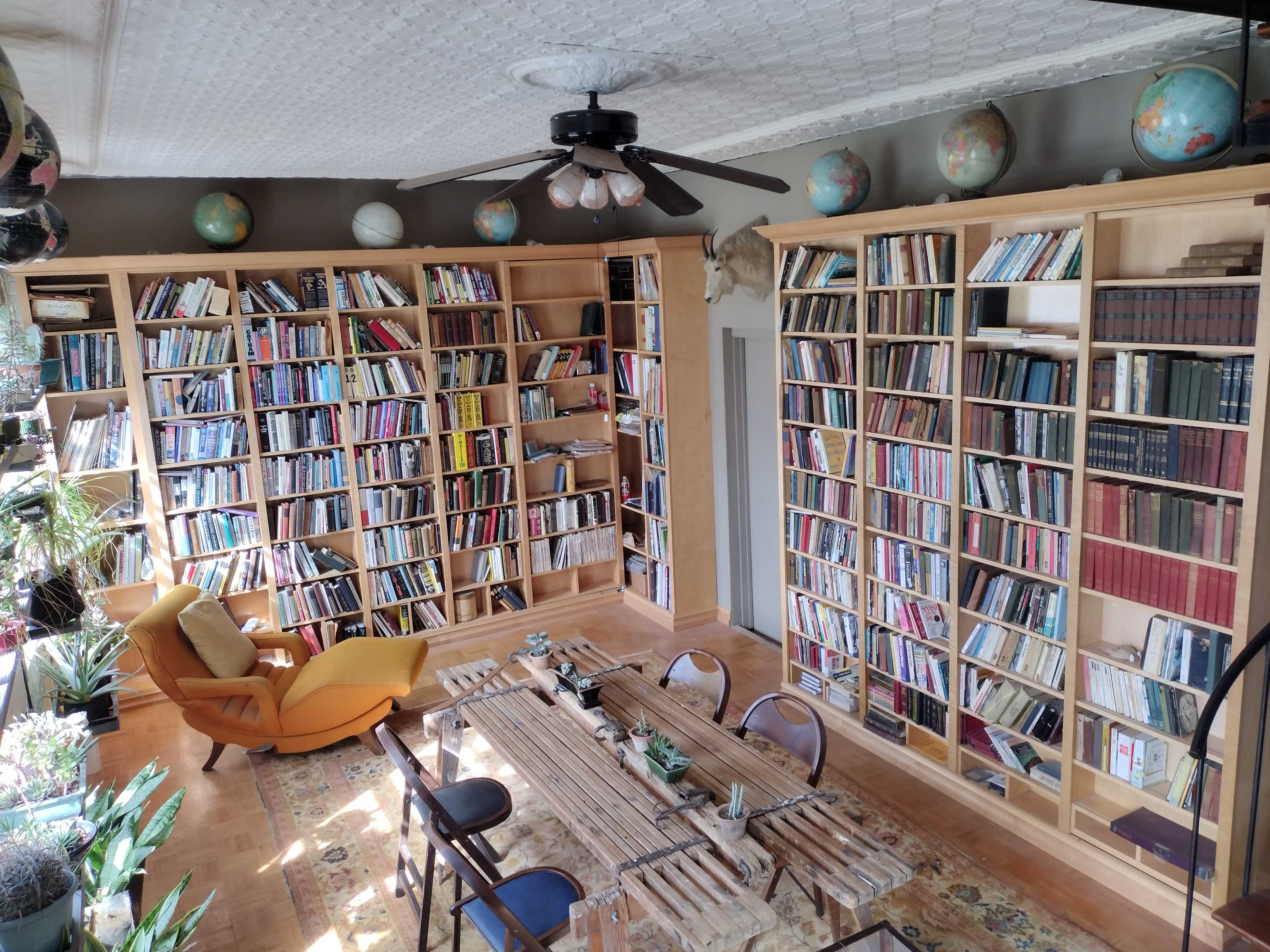 A well-organized library with tall bookshelves filled with books, a central wooden table, and plants around the space.