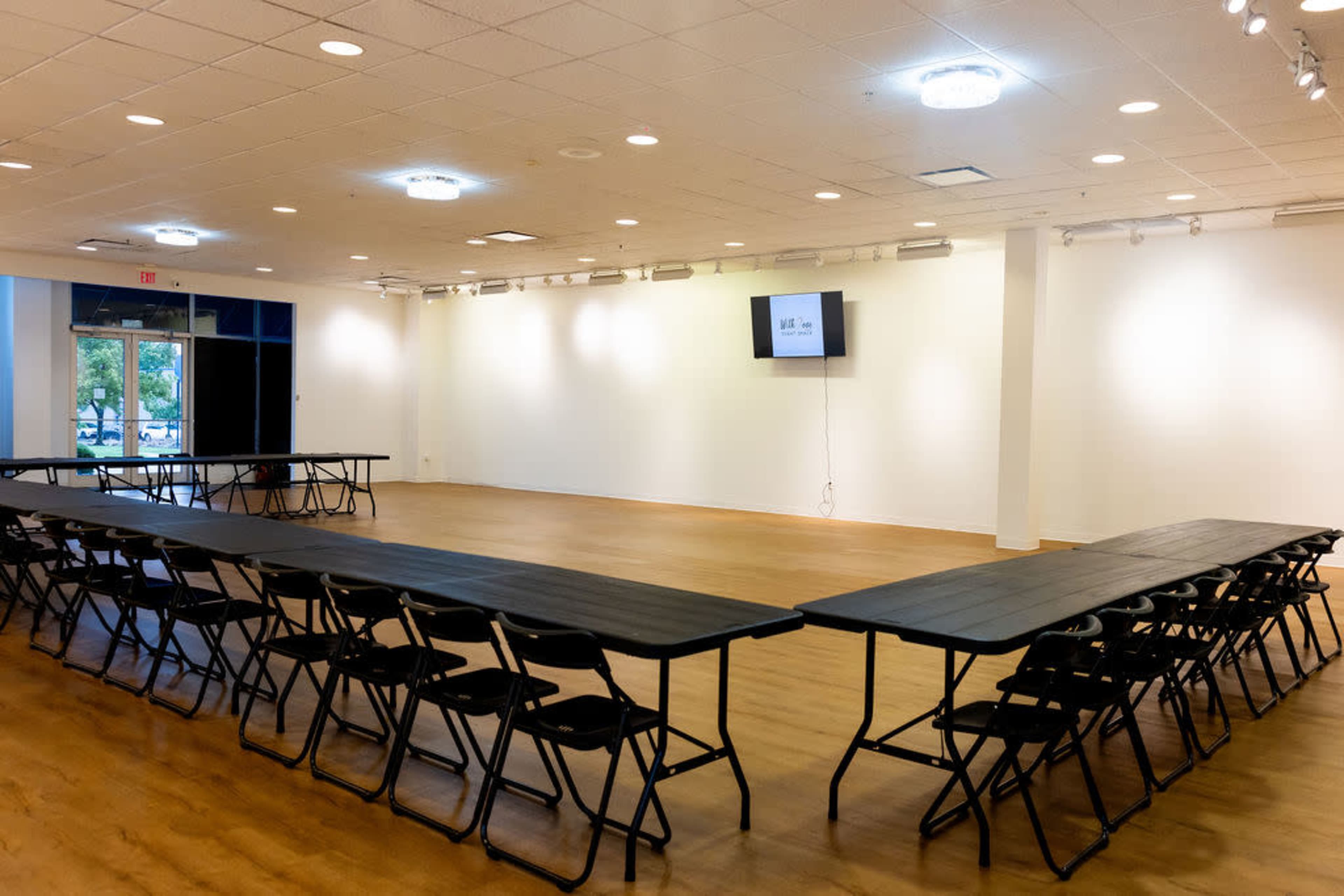 A spacious, well-lit room features two sets of long tables arranged in a U-shape, with black folding chairs placed around them and a screen mounted on the wall.
