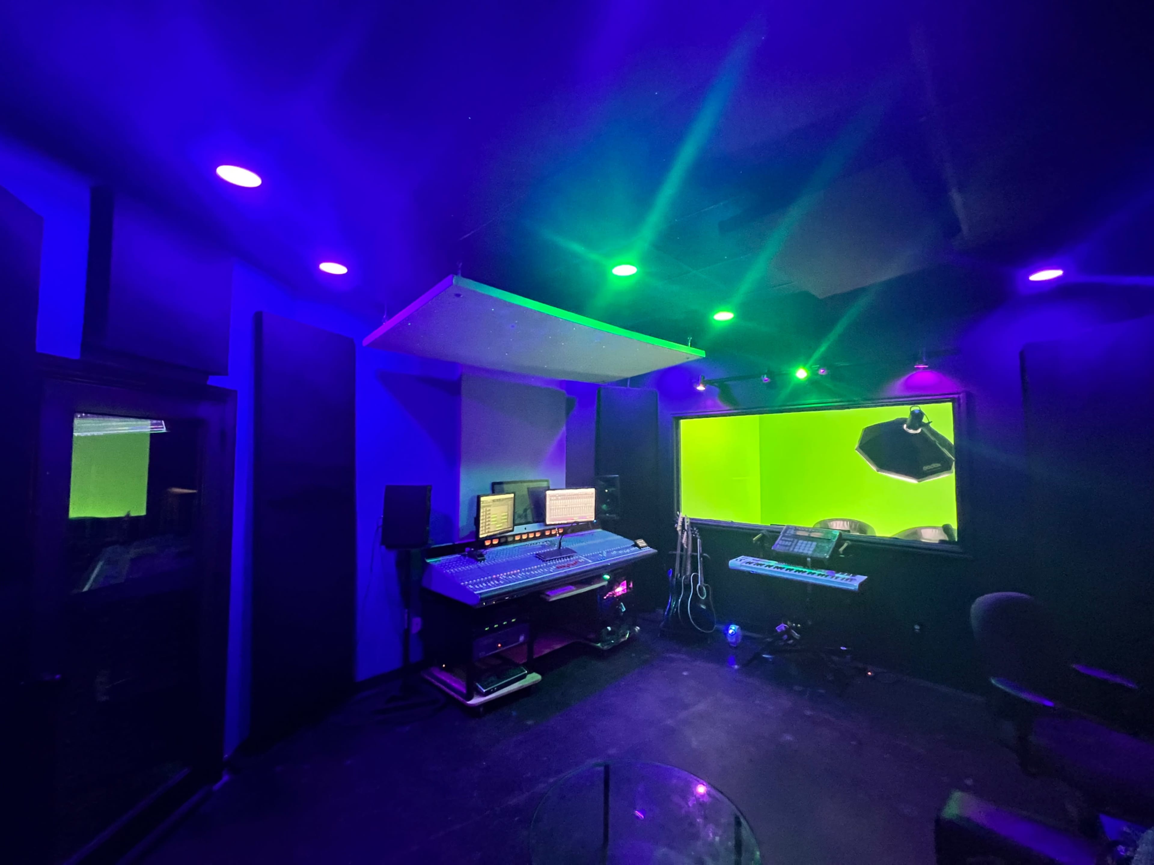 A modern recording studio with a large mixing console, computer monitors, and a vibrant green screen backdrop.
