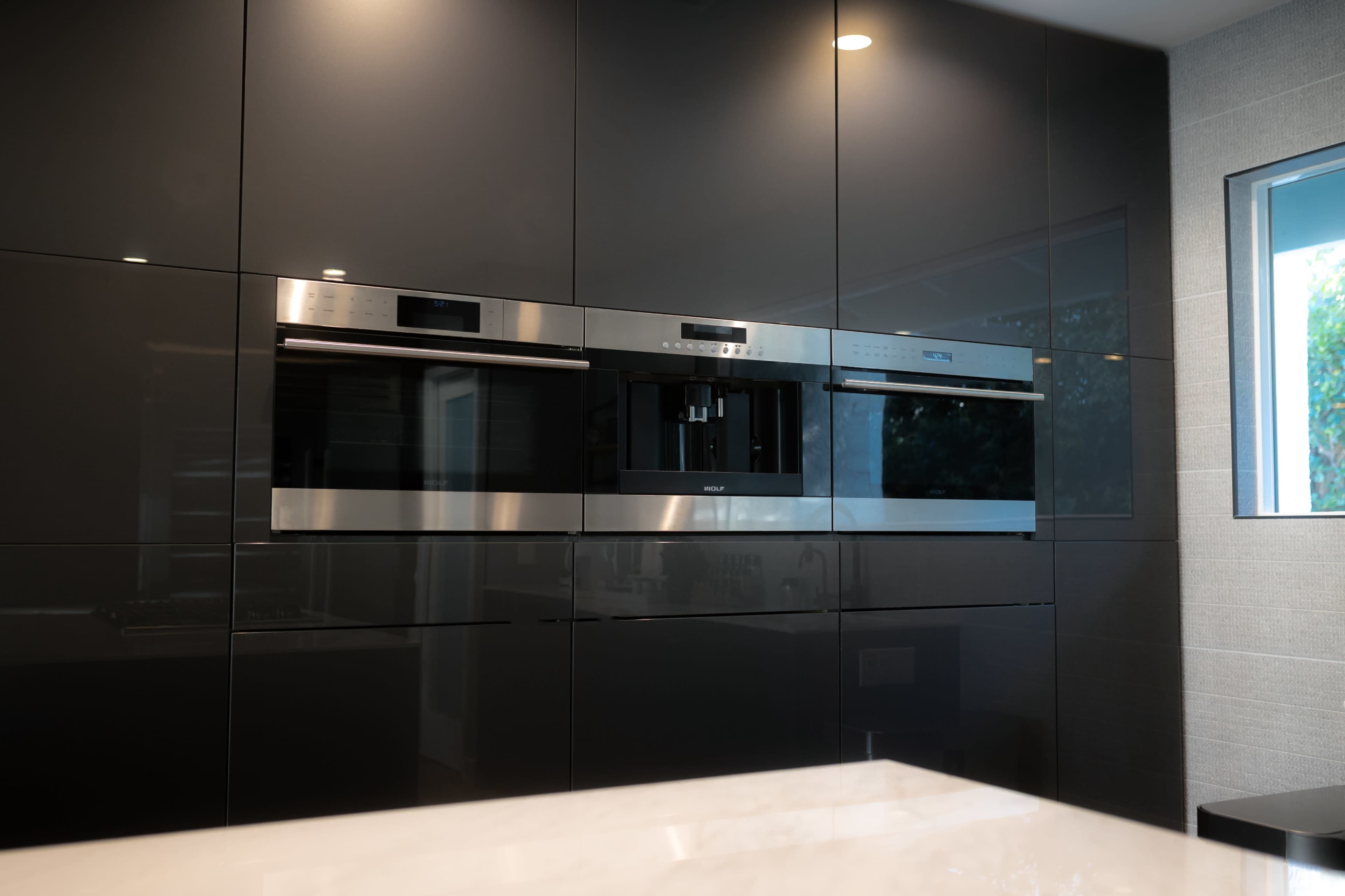 A modern kitchen wall with three built-in stainless steel appliances set against glossy black cabinetry.