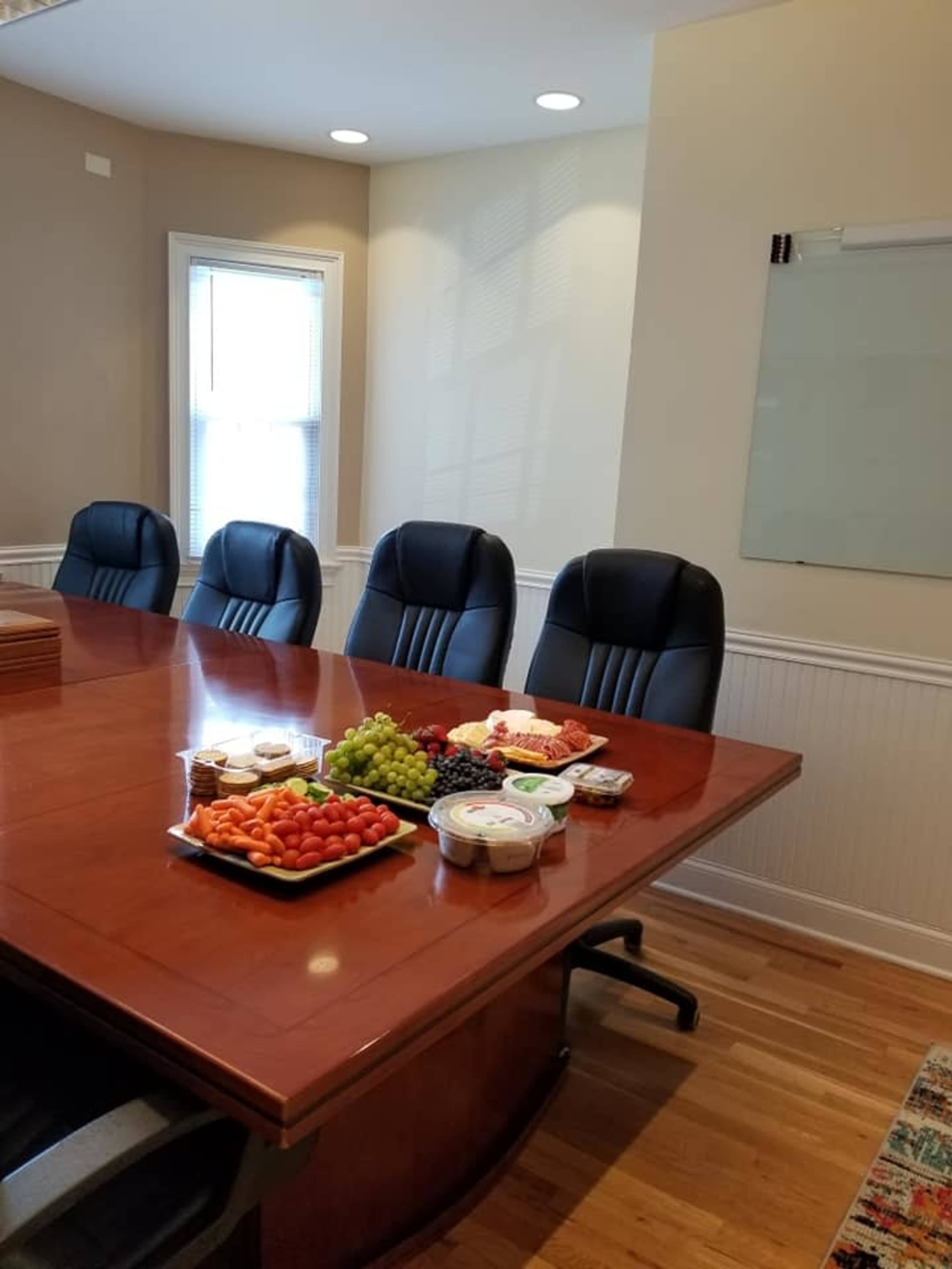 A conference room is set up with a large wooden table displaying an assortment of fruits, vegetables, and snacks, while several black leather chairs are arranged around it.