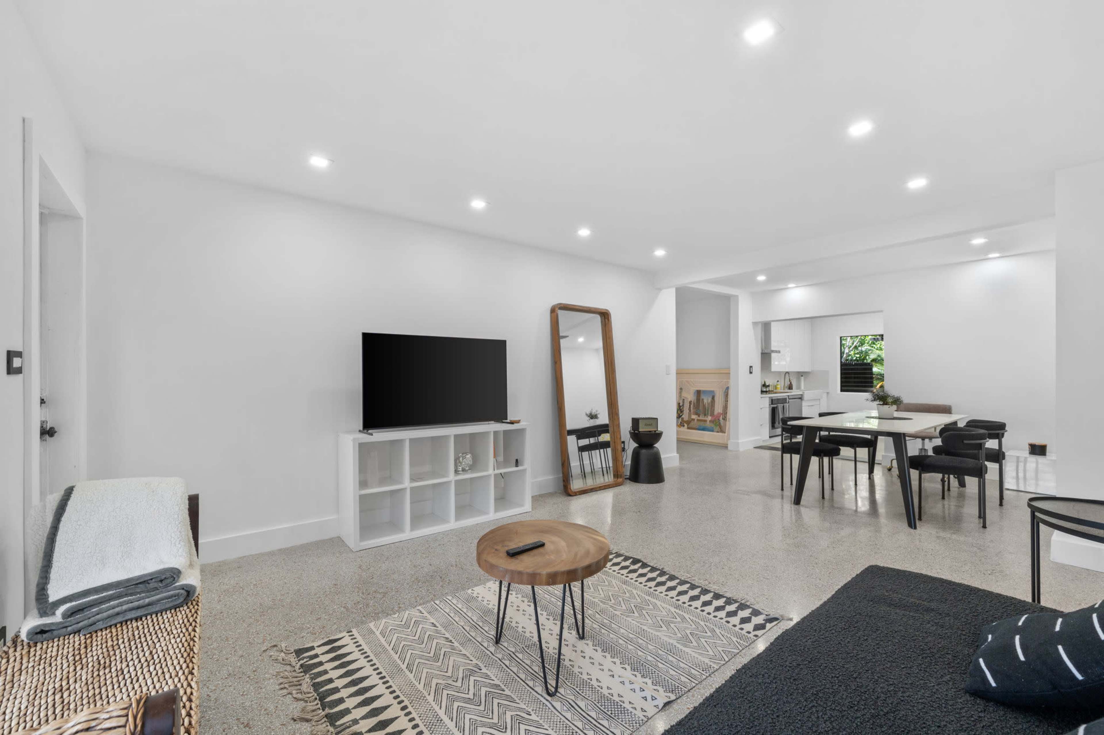 A modern living space with a television, a large mirror, a dining table, and minimalist decor on a polished concrete floor.