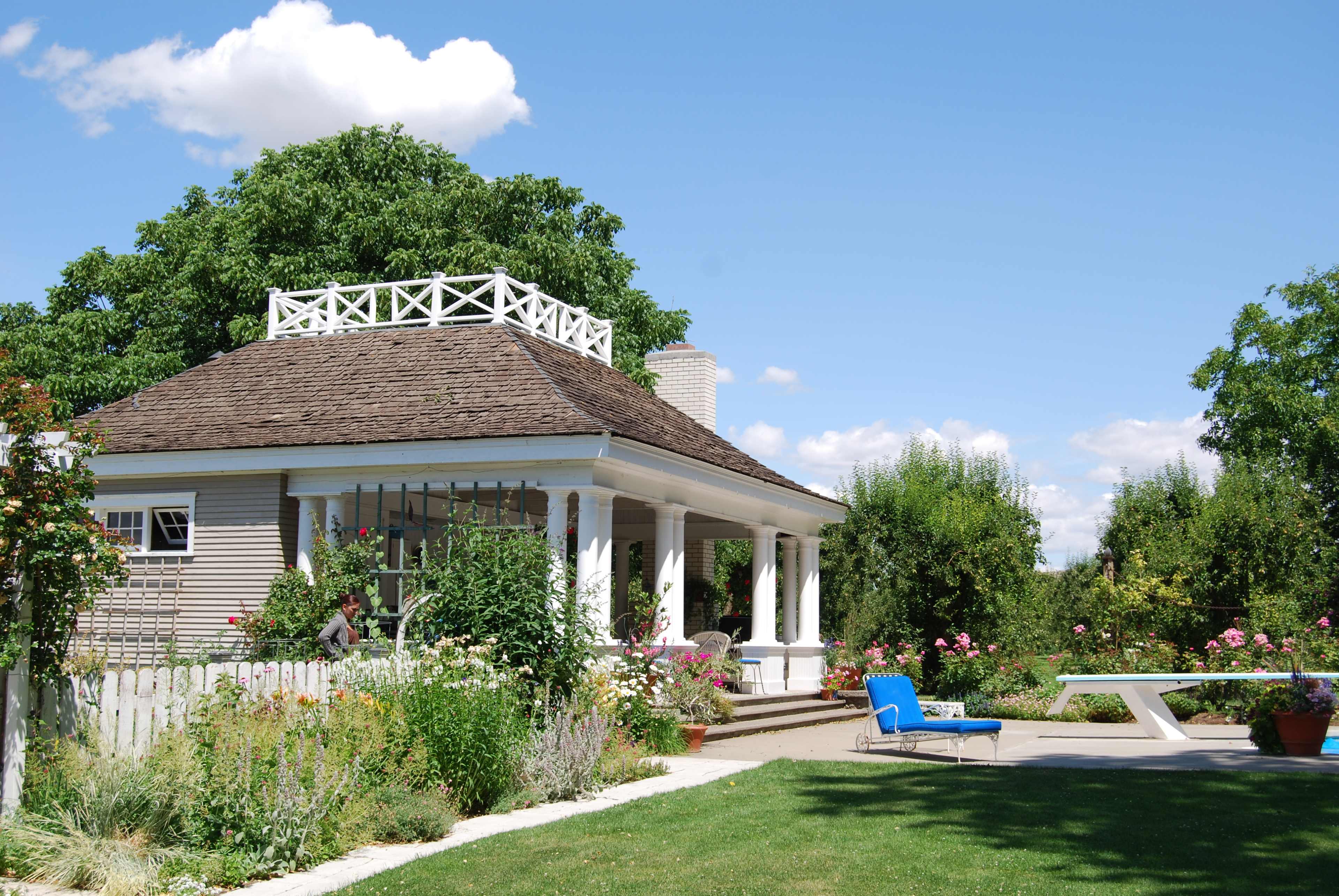 A quaint, single-story house with a round, shingled roof and a balcony is surrounded by colorful flower beds and a well-maintained lawn.