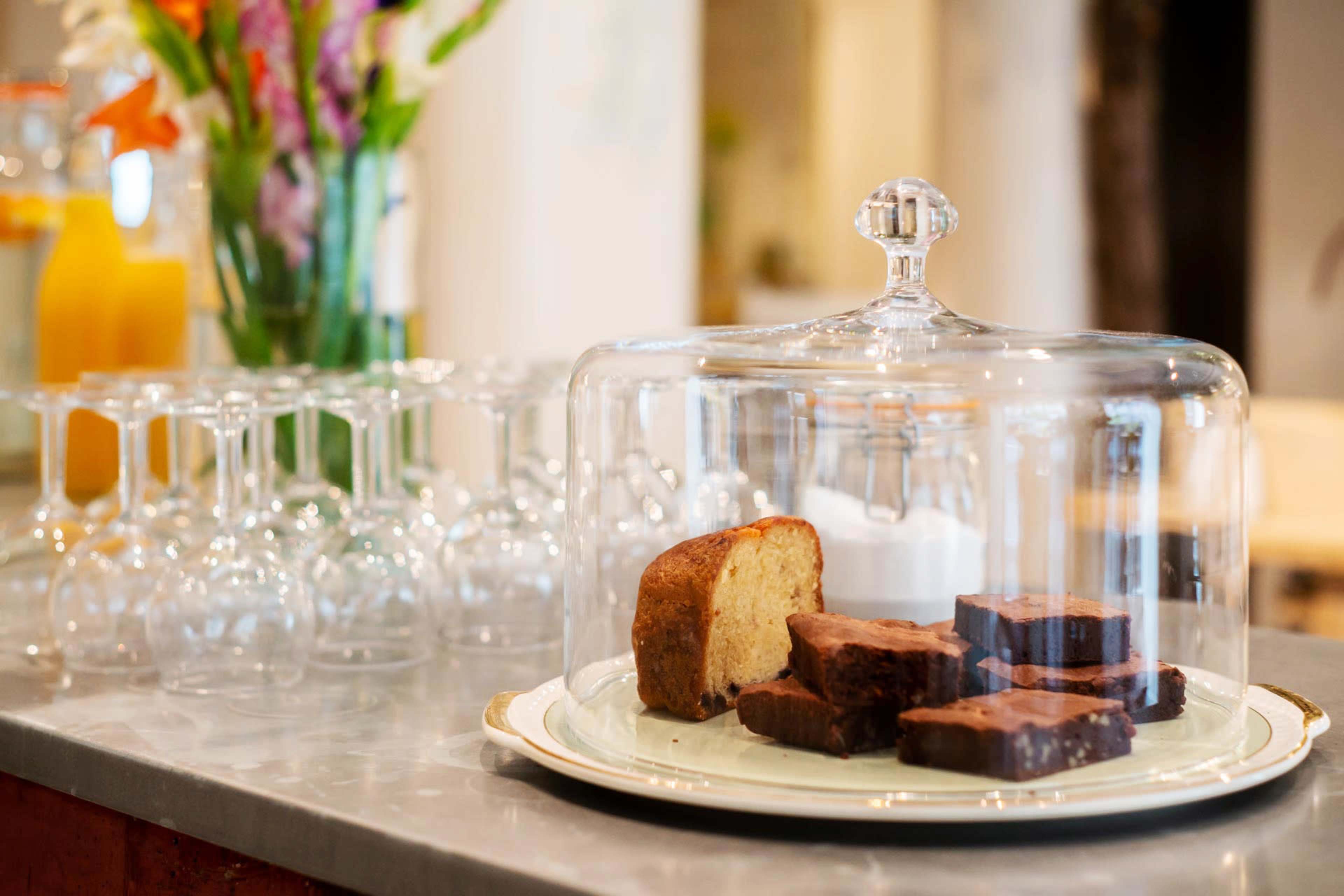 A glass cloche covers a plate of brownies and a slice of cake on a table beside a stack of wine glasses and a bouquet of flowers.