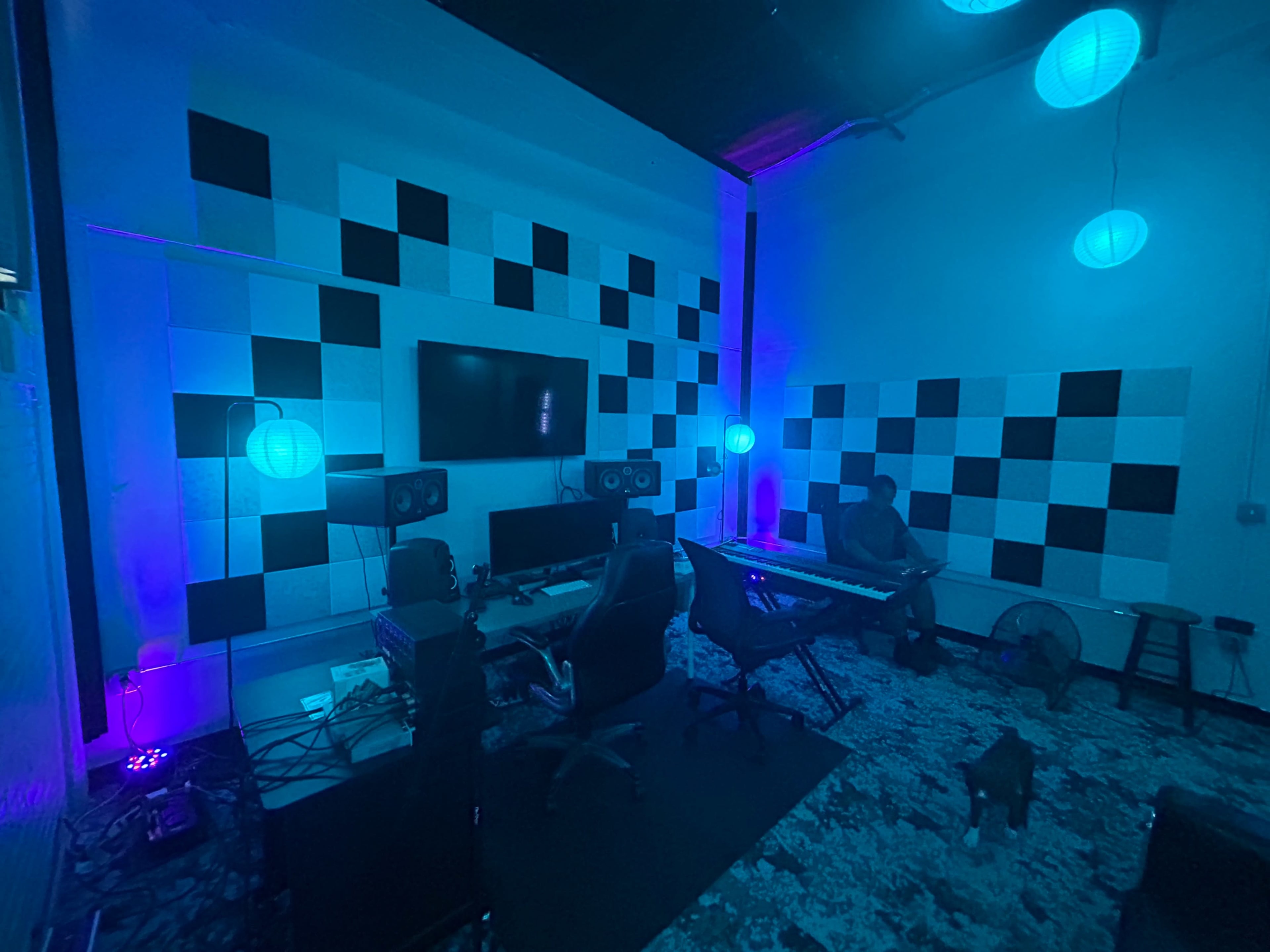 The image shows a music studio featuring a keyboard, multiple monitors, and colorful ambient lighting with a person seated at a workstation.