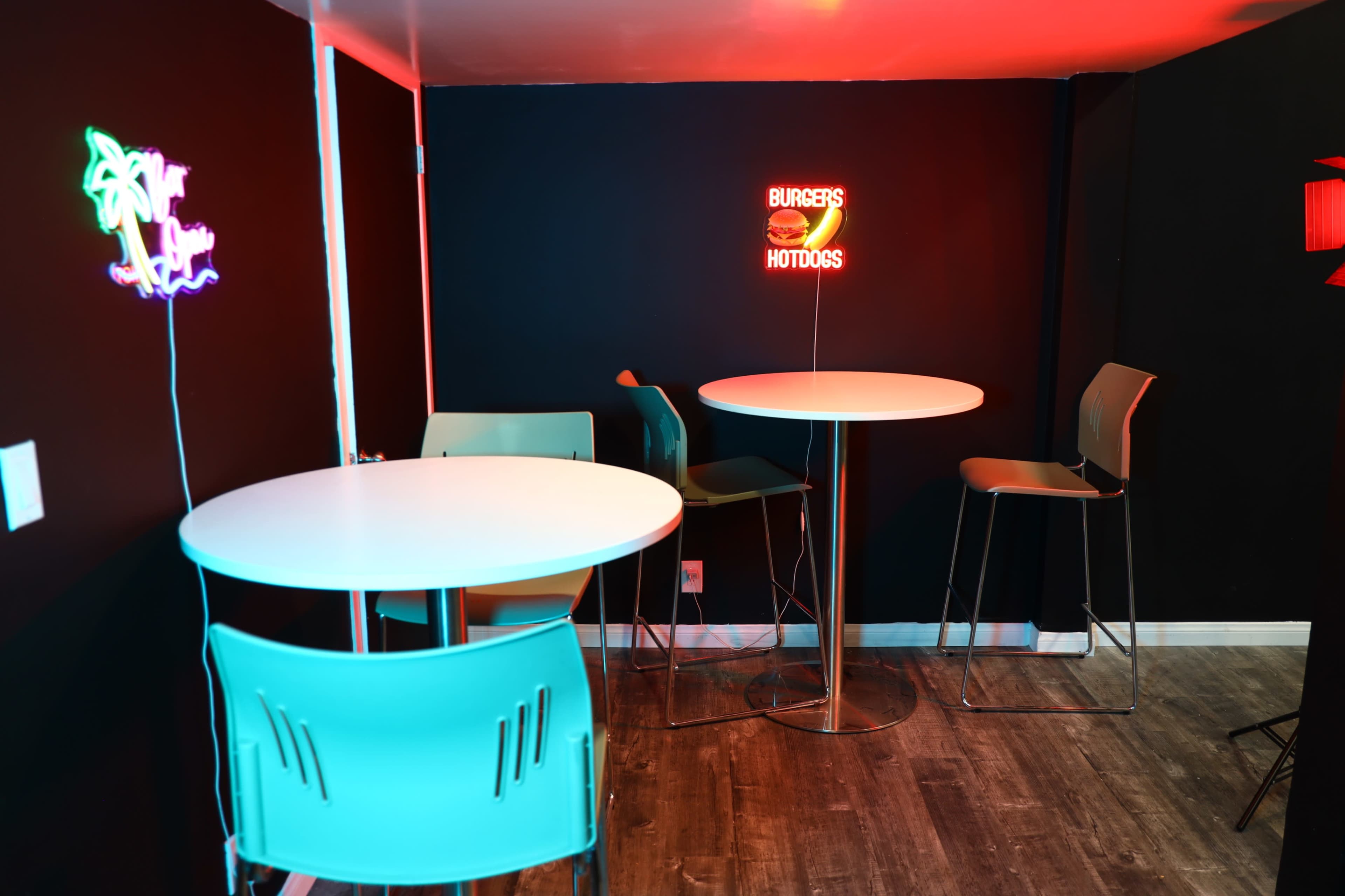 The image shows a small indoor space with two high tables, modern chairs, and neon signs that read "BURGERS" and "HOTDOGS."