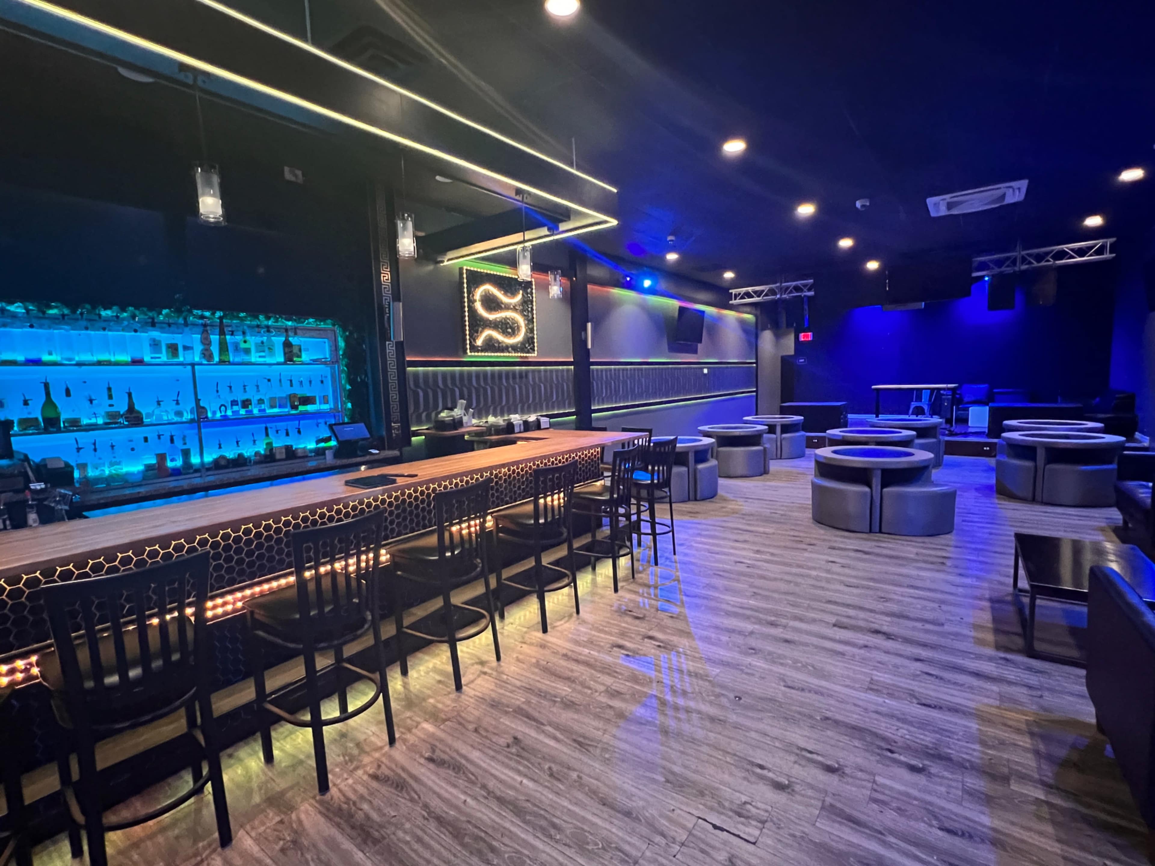 A dimly lit bar area with a blue-lit backlit shelf displaying various bottles, surrounded by seating arrangements of circular booths and chairs.