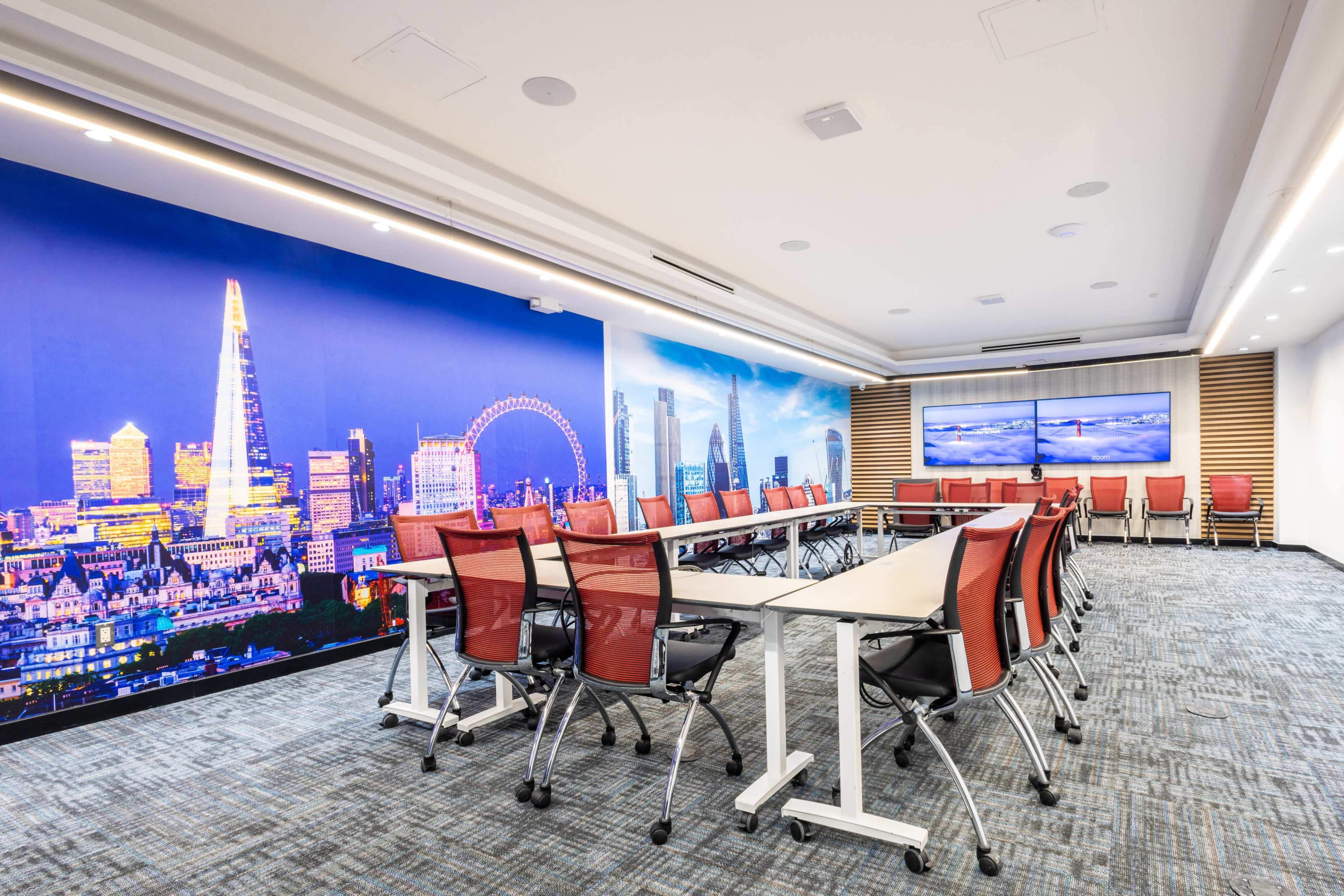 The image shows a modern conference room with a long table surrounded by red chairs and a wall featuring a large city skyline backdrop.