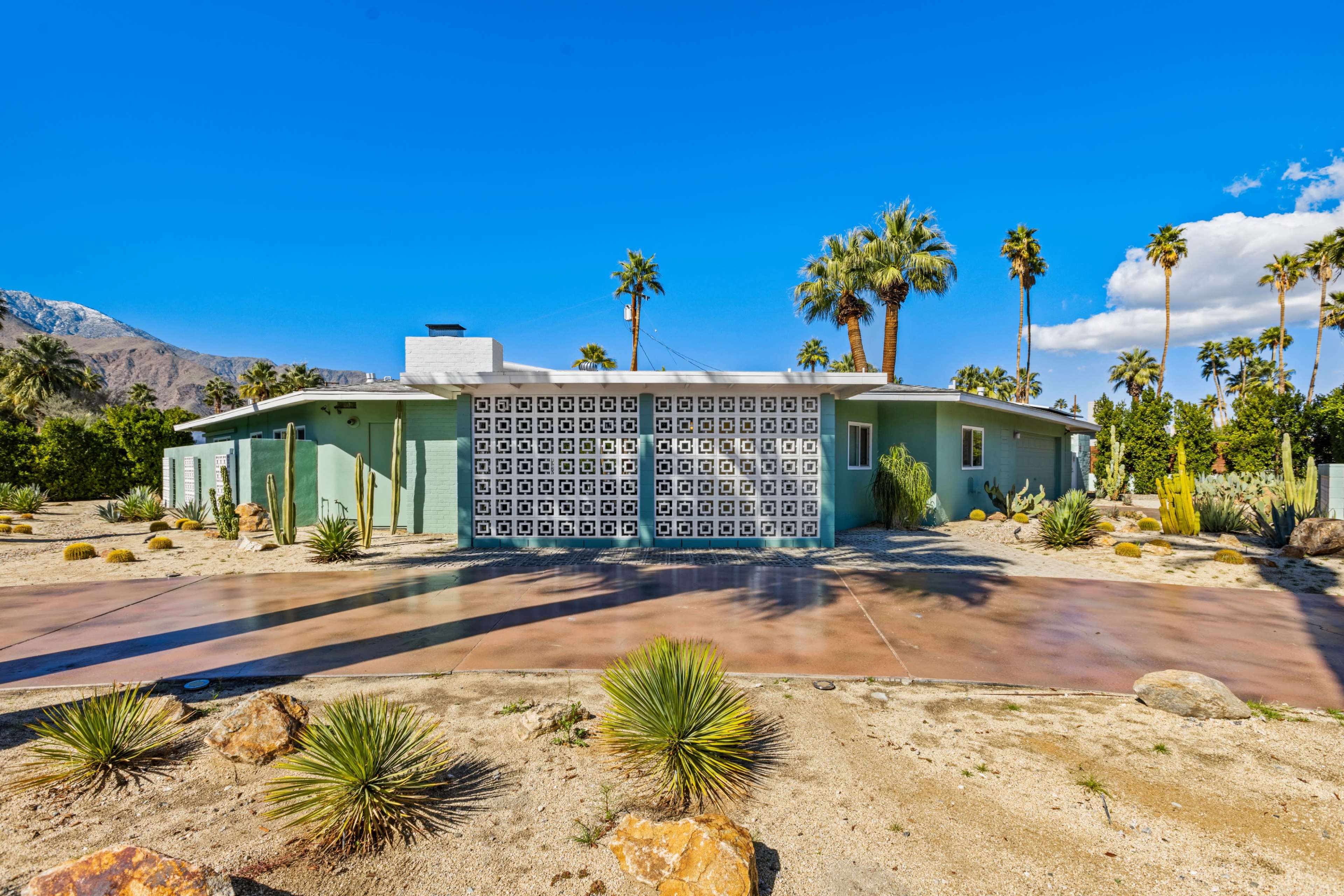 A single-story house with a decorative block wall facade is surrounded by desert landscaping and palm trees.