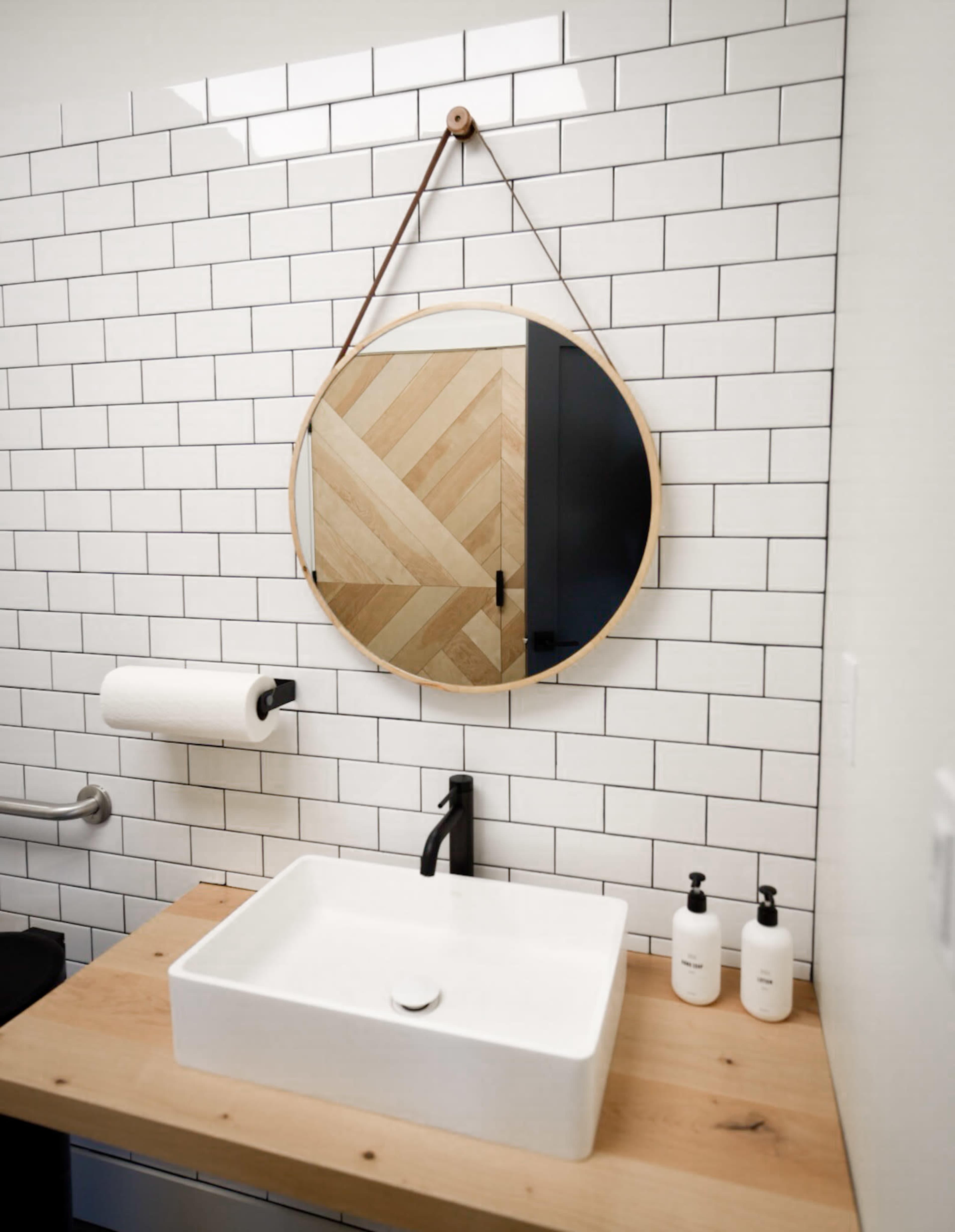 A circular mirror with a wooden frame hangs above a white rectangular sink on a wooden countertop in a bathroom with white subway tile walls.