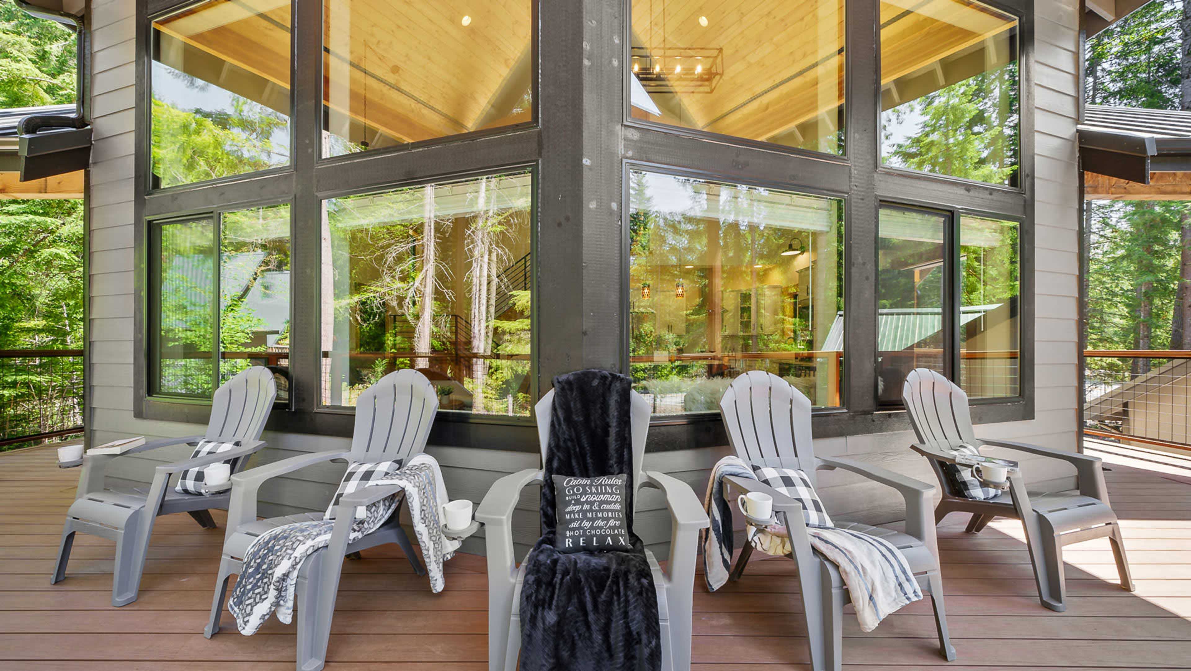 A seating area with four gray adirondack chairs, each accompanied by a small table and a throw blanket, is positioned on a wooden deck surrounded by large windows and trees.