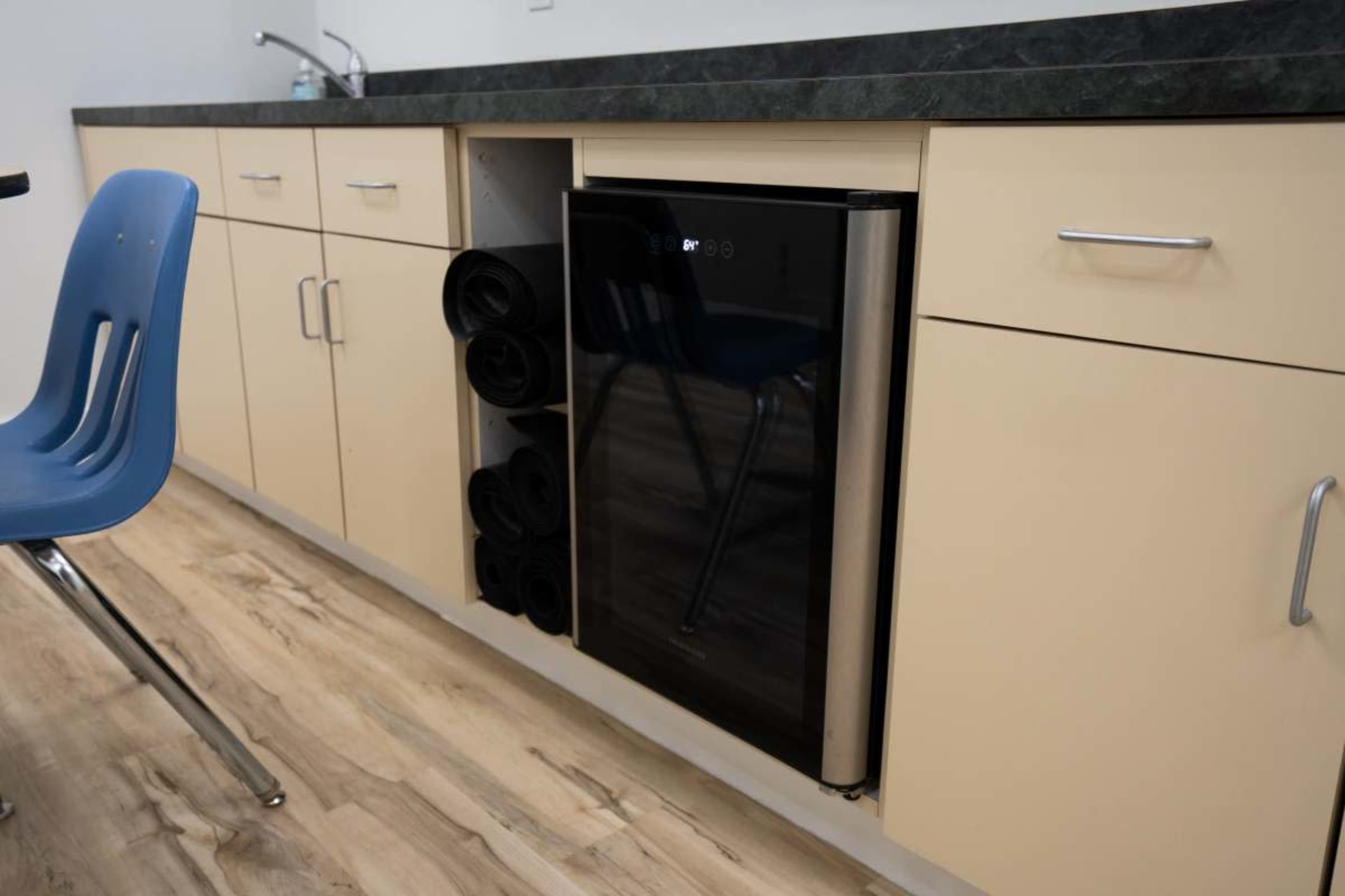 A kitchen countertop features a compact refrigerator alongside neatly rolled towels stored in a cabinet.