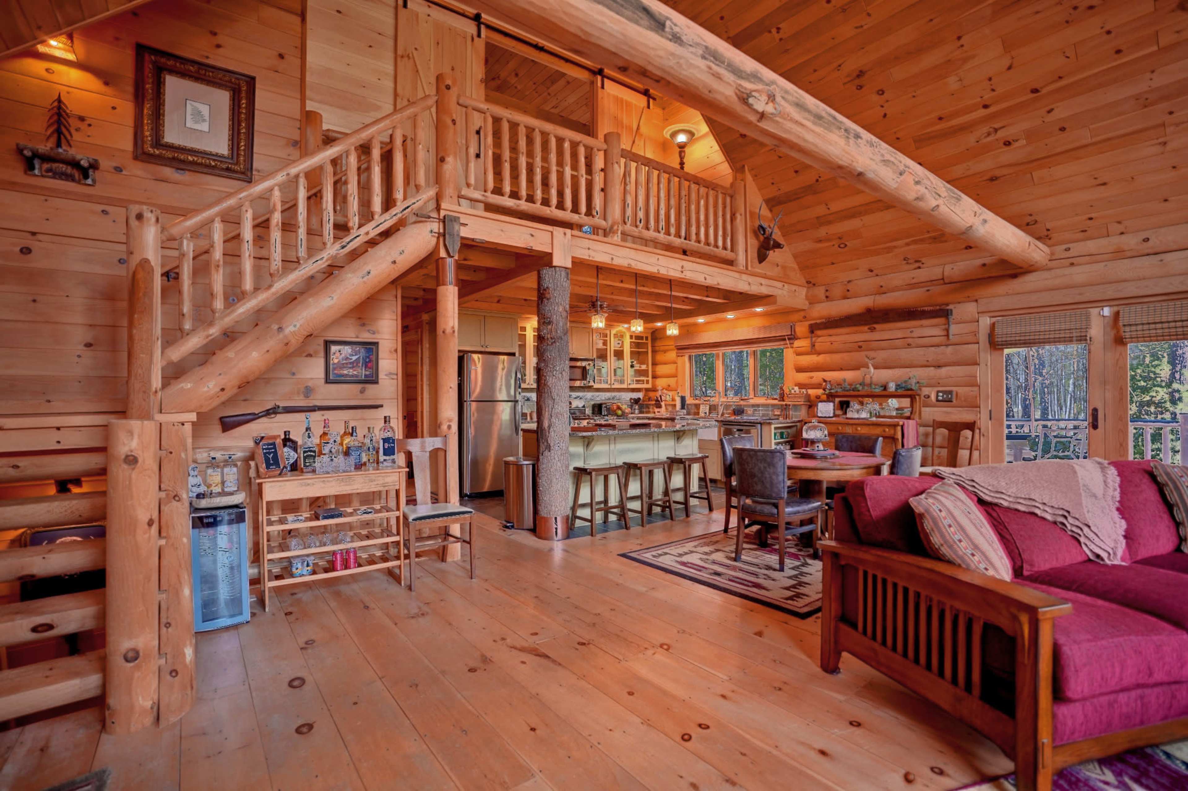 The image shows a cozy, wooden interior of a log cabin featuring a living area, kitchen, and a loft space connected by a staircase.