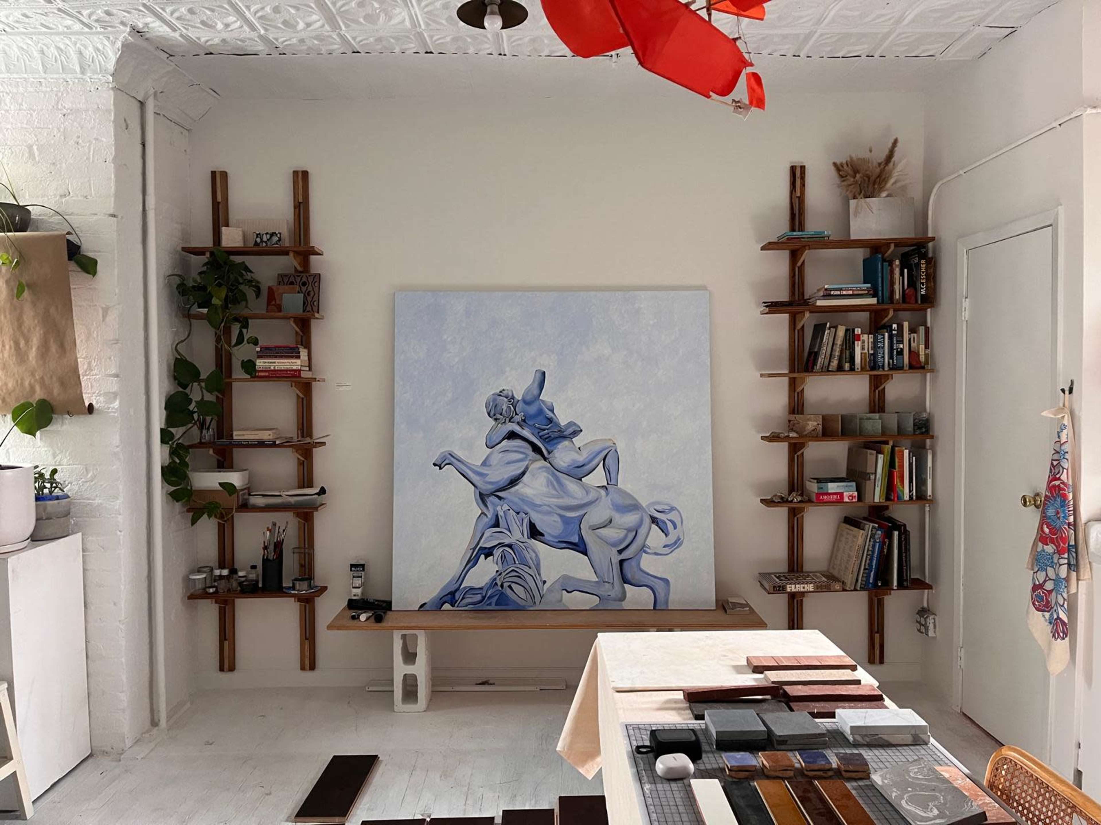 The image depicts an art studio with wooden shelves displaying books and plants, a large blue-toned painting of a figure on a horse, and a table with various materials arranged on it.