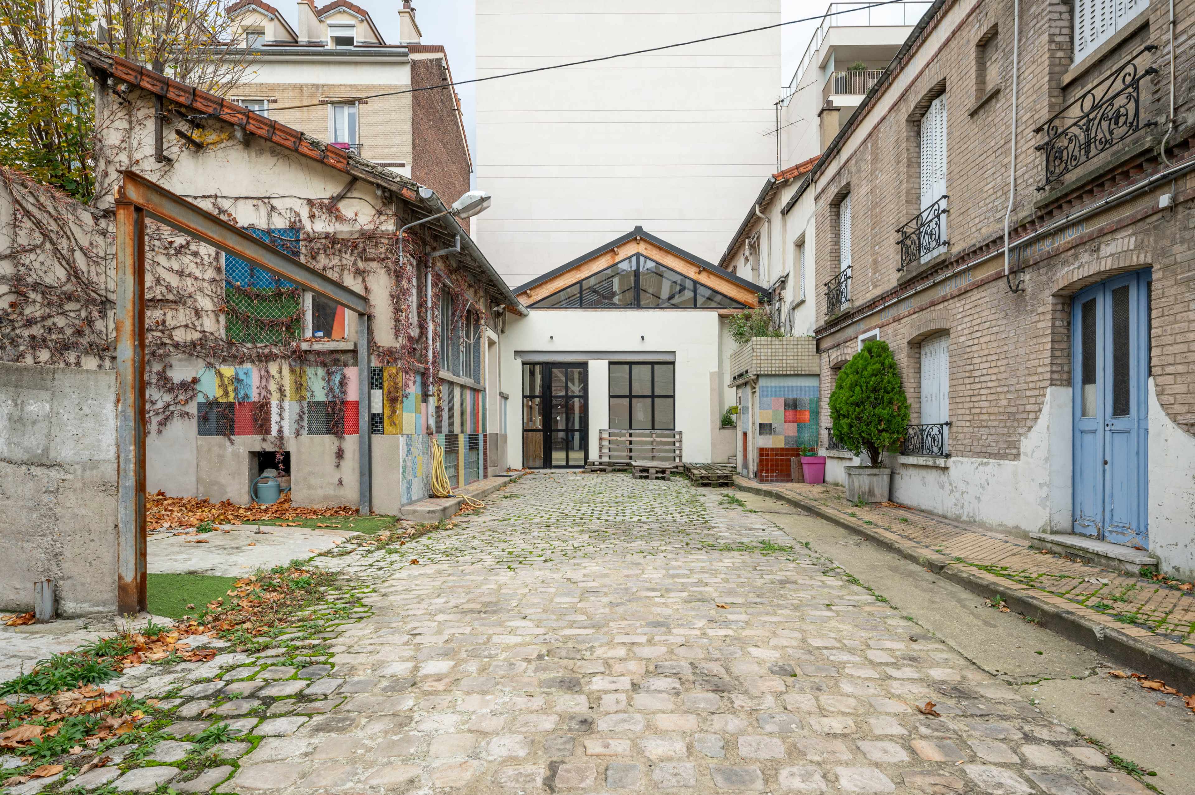 A cobblestone pathway leading to a modern building framed by two older brick structures, with a mix of greenery and colorful, patterned walls.