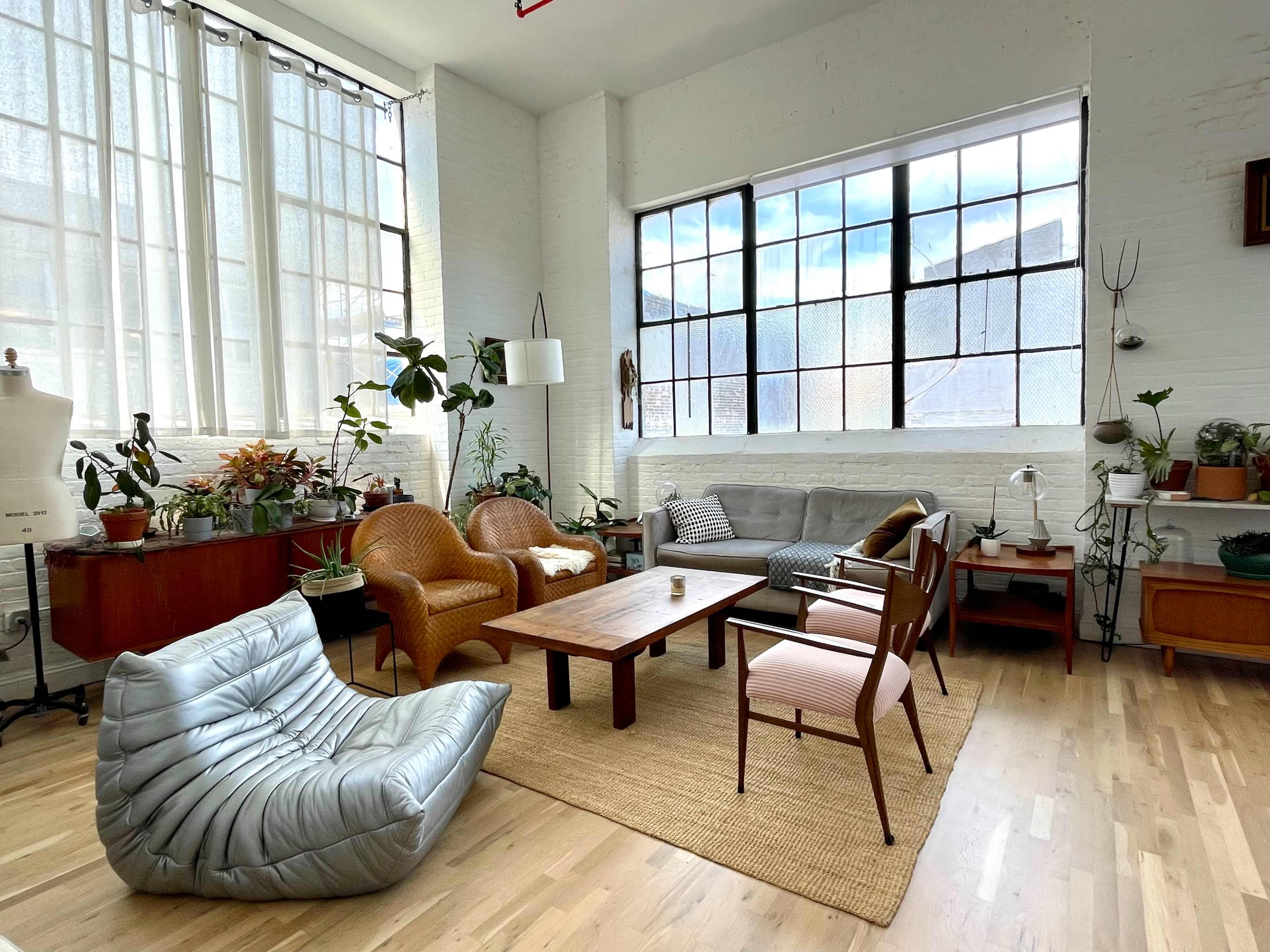 The image shows a bright, spacious living area with a mix of seating options, including a grey sofa, orange armchairs, and a unique beanbag, surrounded by indoor plants and natural light from large windows.