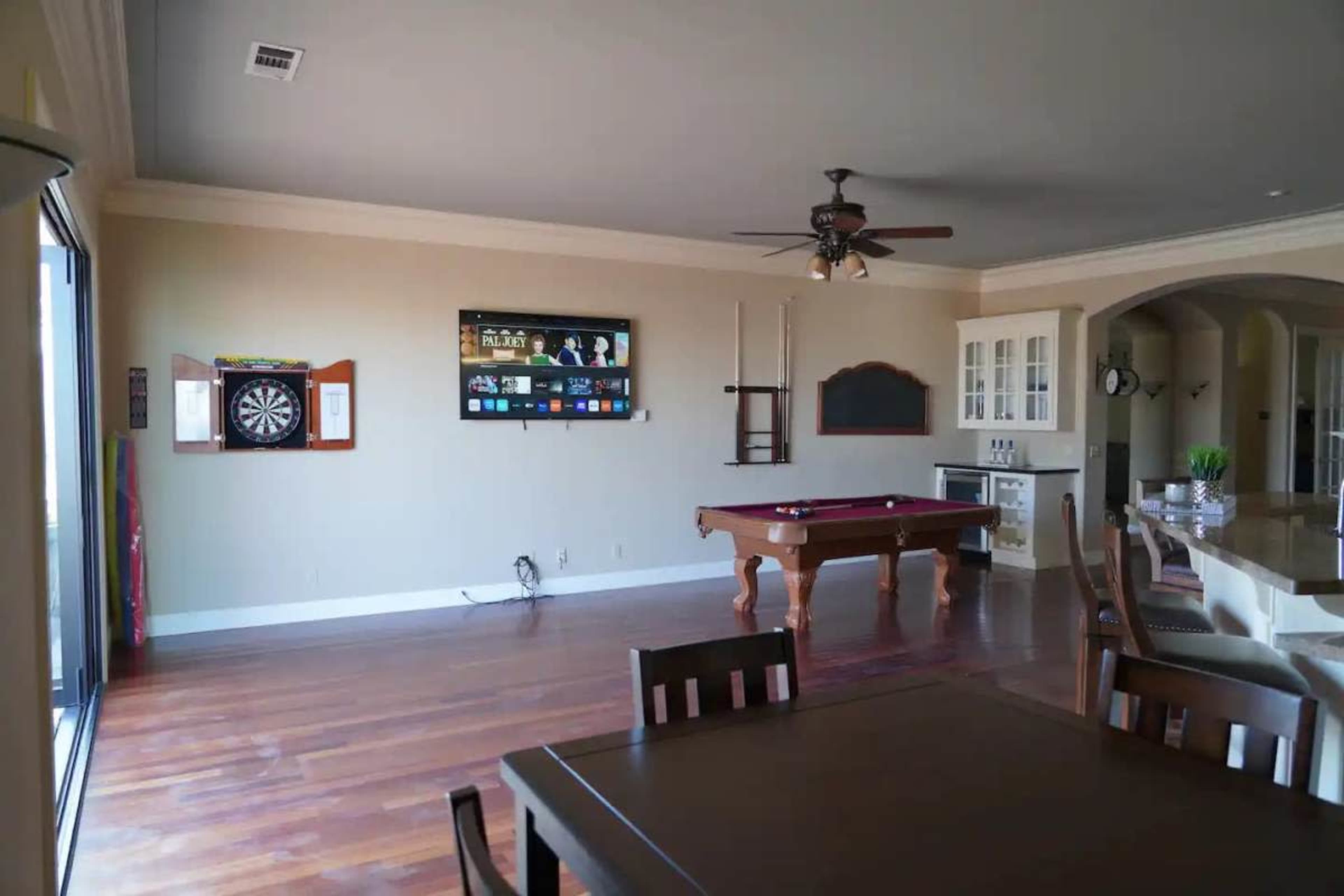 The image shows a spacious game room with a pool table, a dartboard on the wall, and a dining area with wooden furniture.