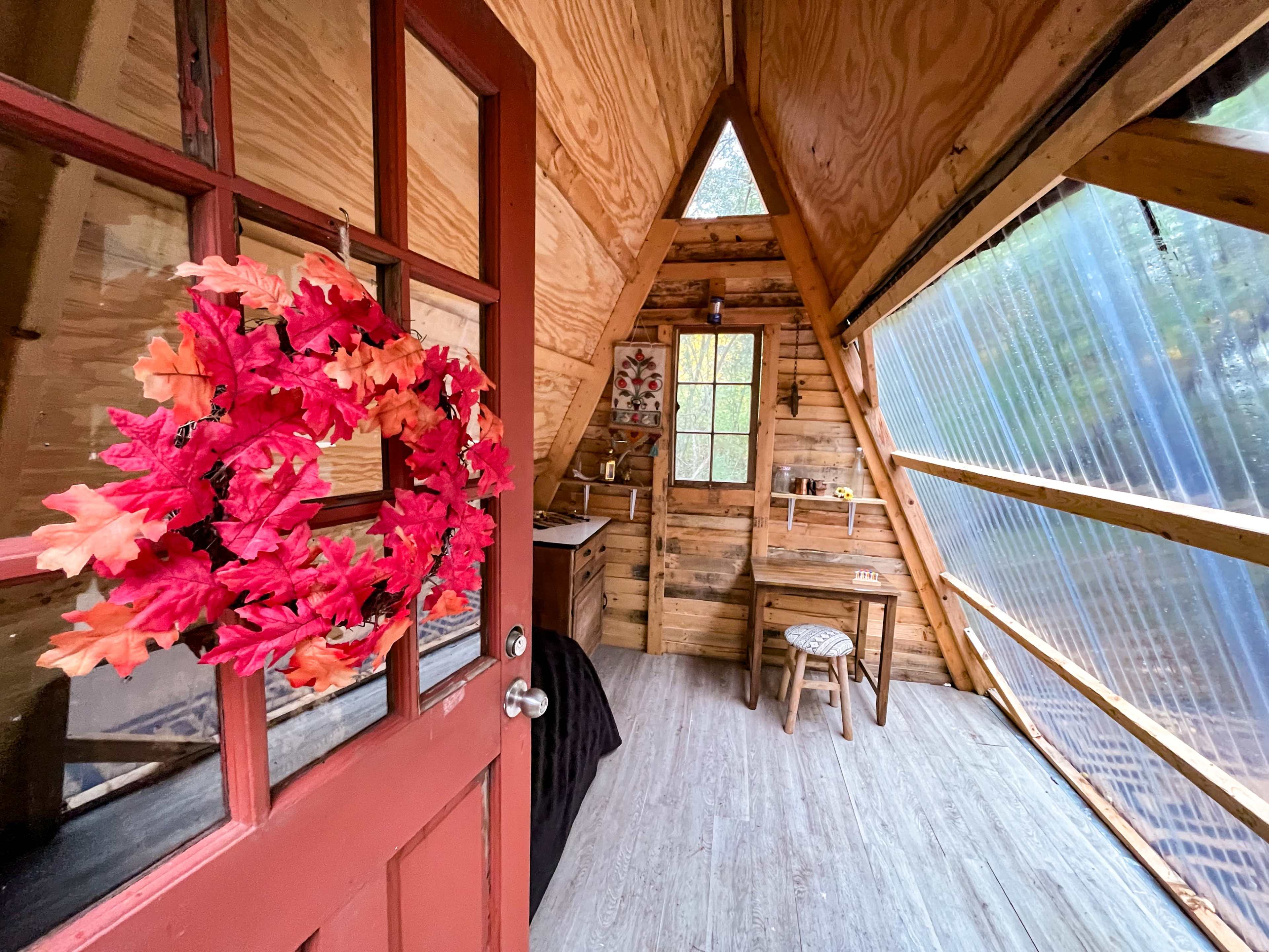 The interior of a wooden A-frame cabin features a door with a floral wreath, a small table, and a large window allowing natural light.