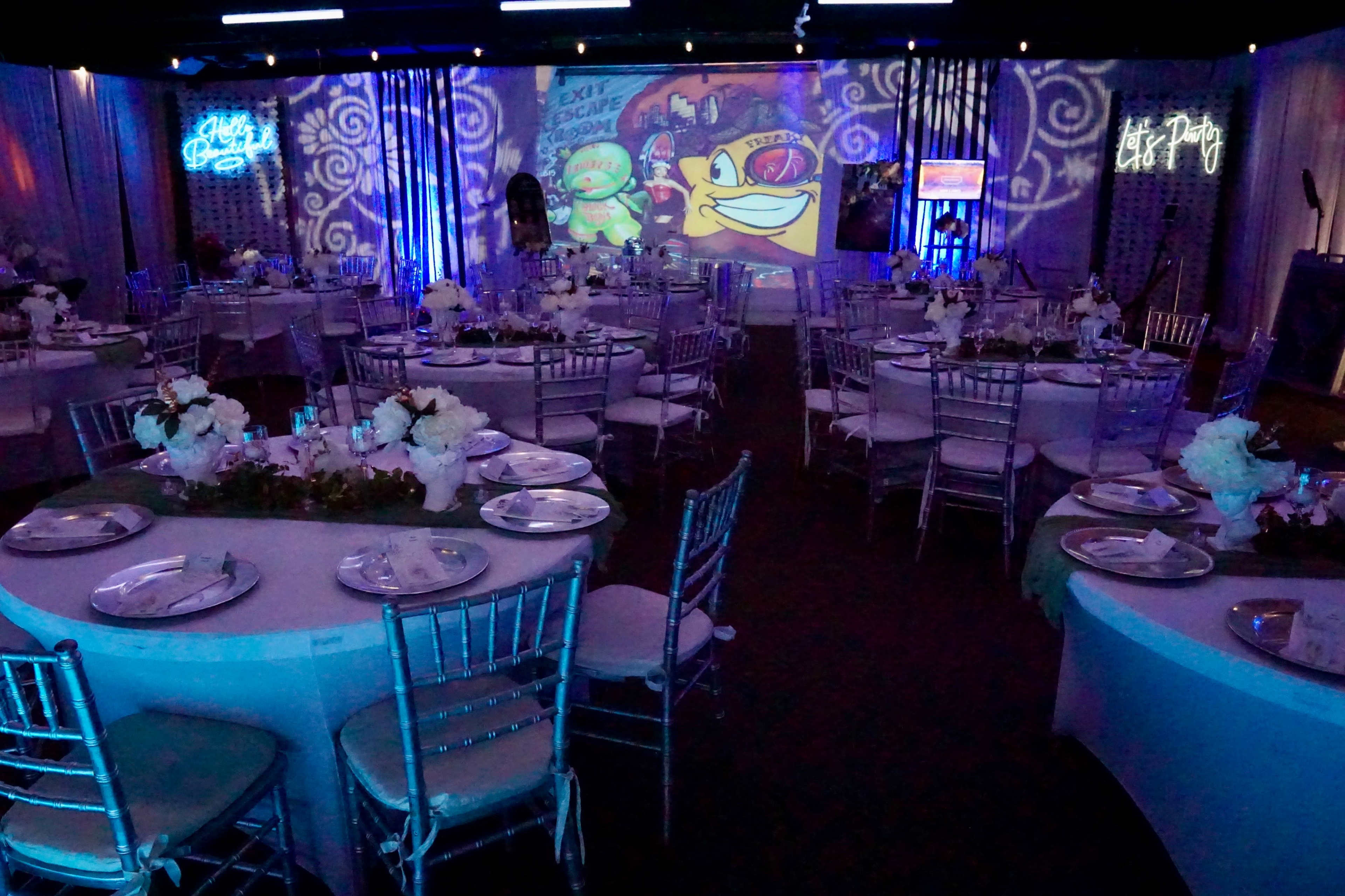 A banquet hall arranged with round tables set for a formal event, featuring floral centerpieces and colorful projected graphics on the walls.