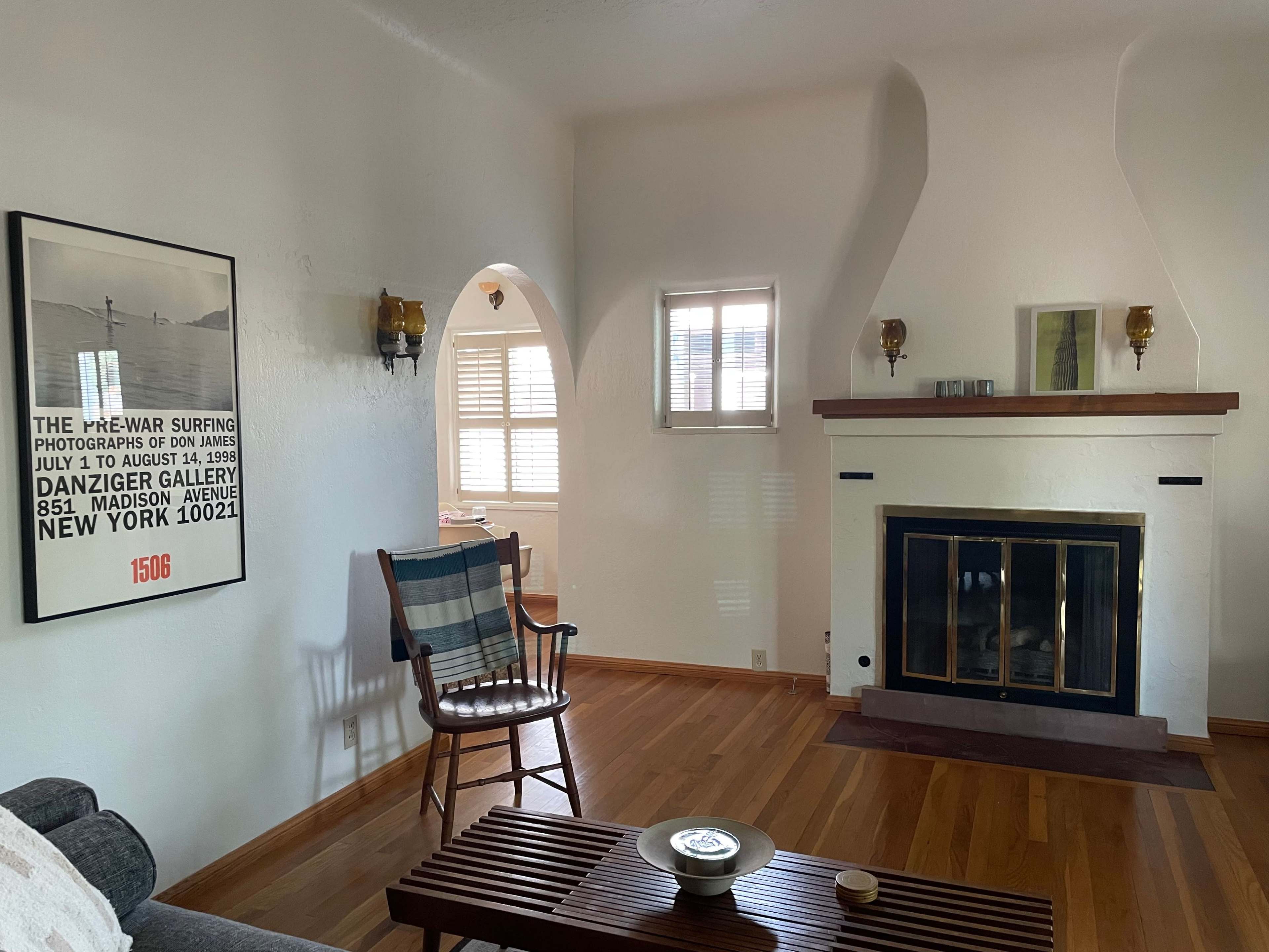 A living room with a wooden floor, a fireplace, a framed poster on the wall, and a chair next to a coffee table.