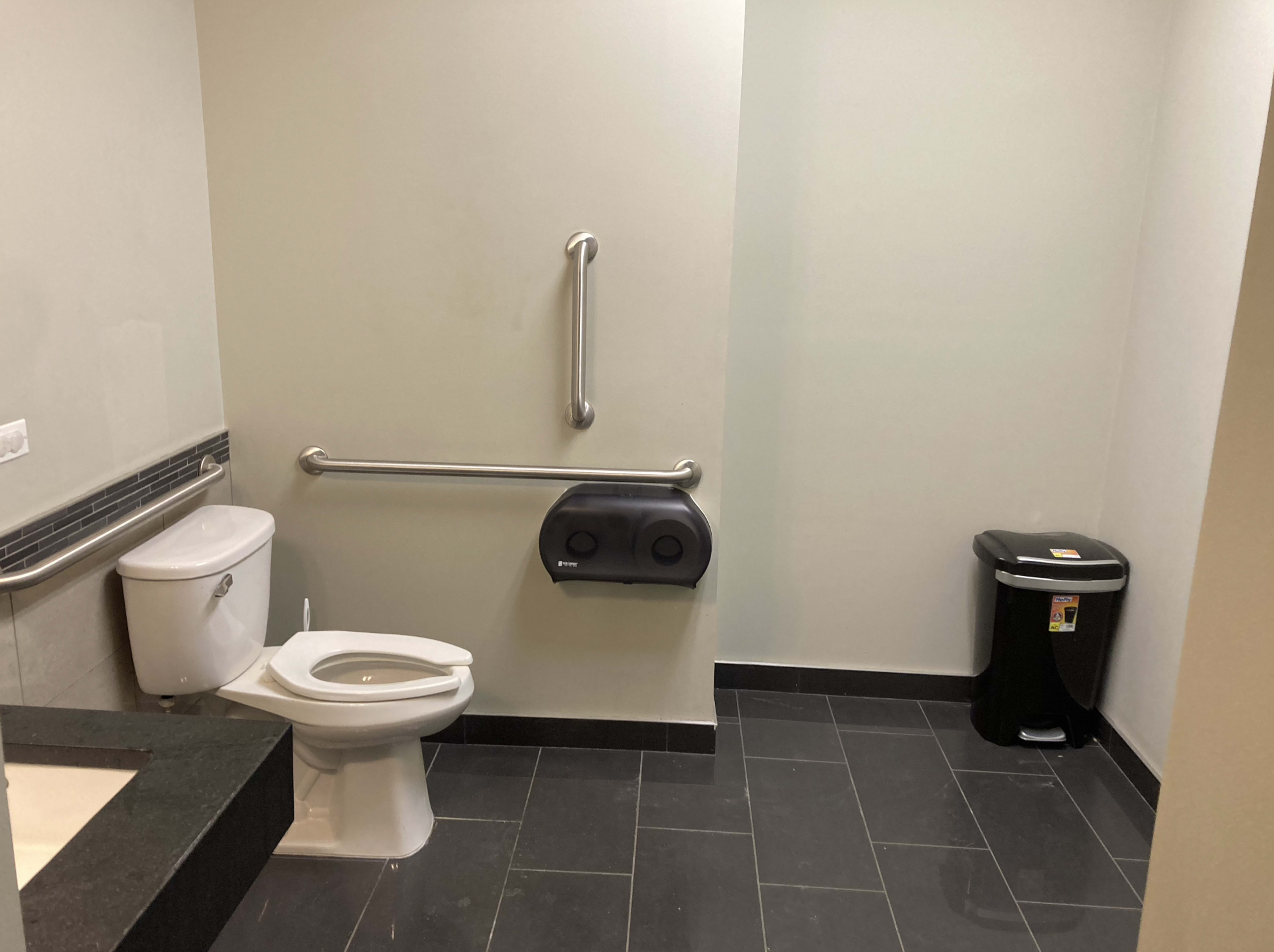 The image shows a clean, simple restroom with a toilet, toilet paper dispenser, grab bar, and a trash can in the corner.