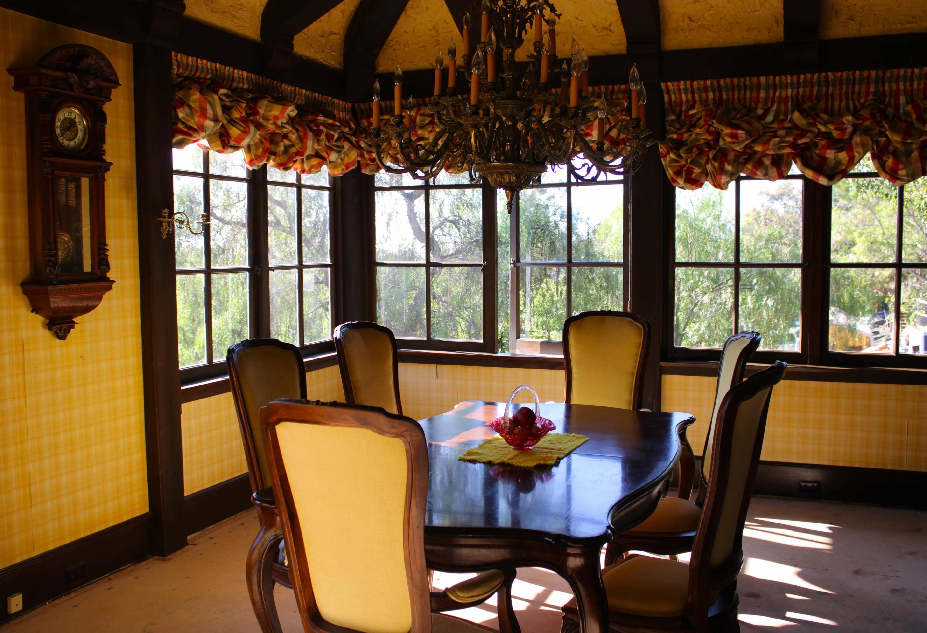 The image shows a dining room with a wooden table, six upholstered chairs, a chandelier, and large windows surrounded by yellow patterned walls and a decorative window valance.
