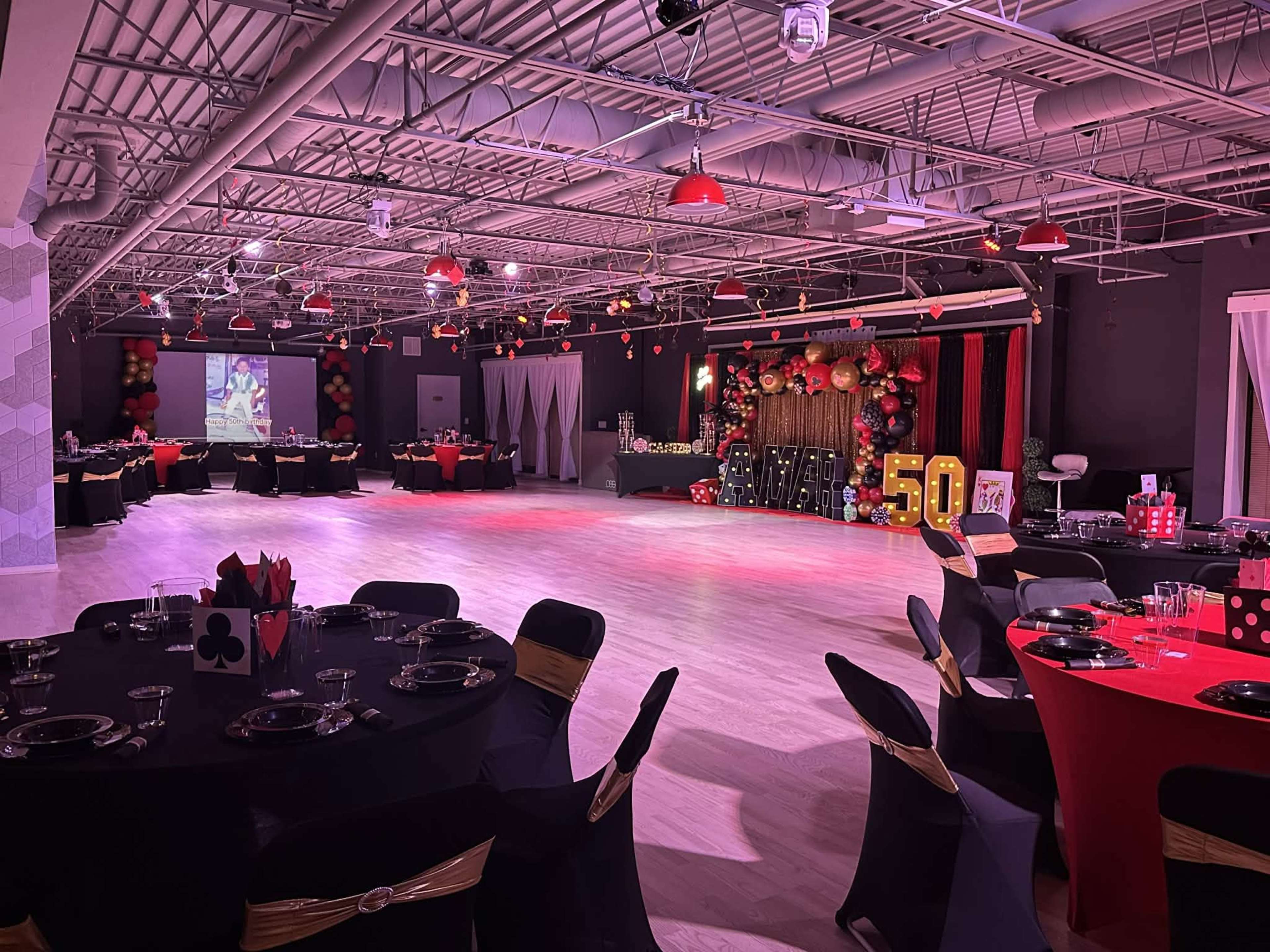 The image shows a spacious event venue set up for a celebration, with tables dressed in black and red linens, and decorative elements including balloons and a backdrop featuring the number 50.