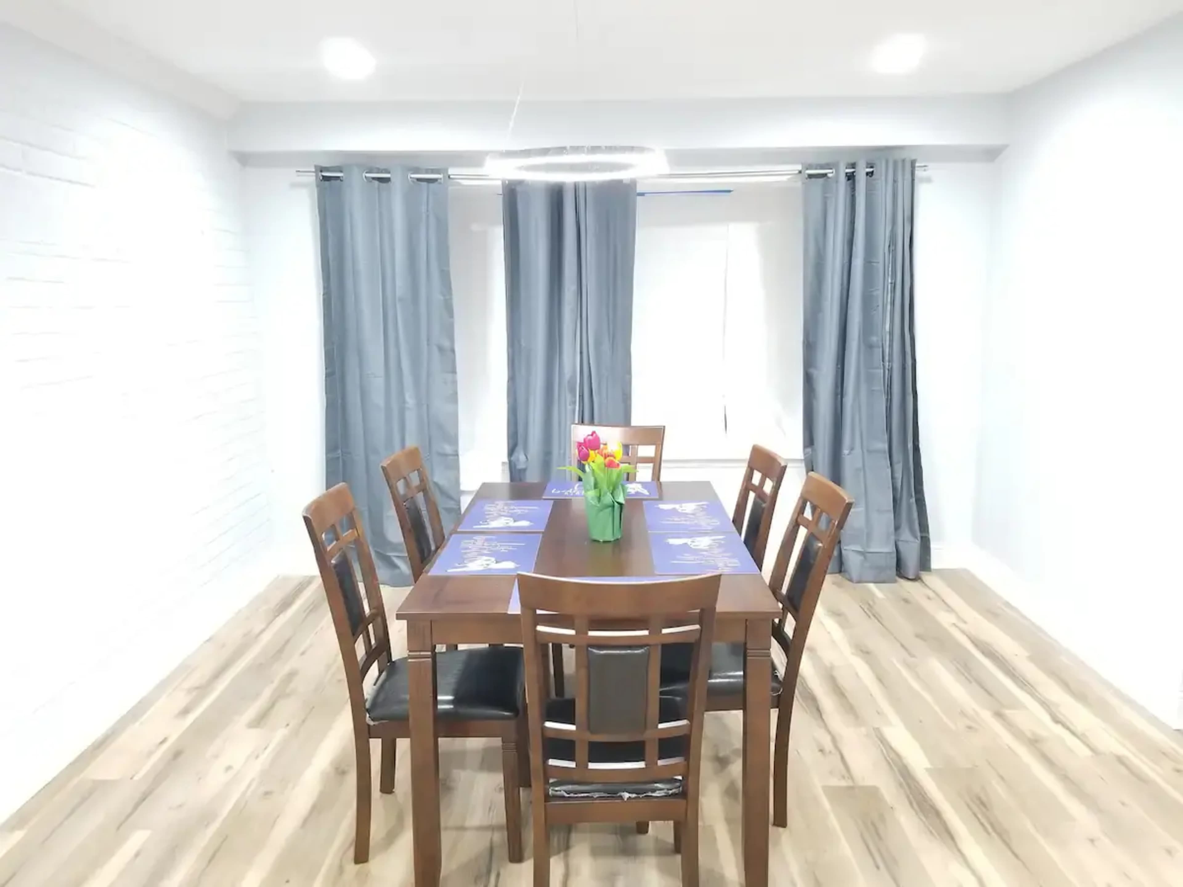 A dining room features a wooden table surrounded by six chairs, with a vase of flowers at the center and gray curtains framing the windows.
