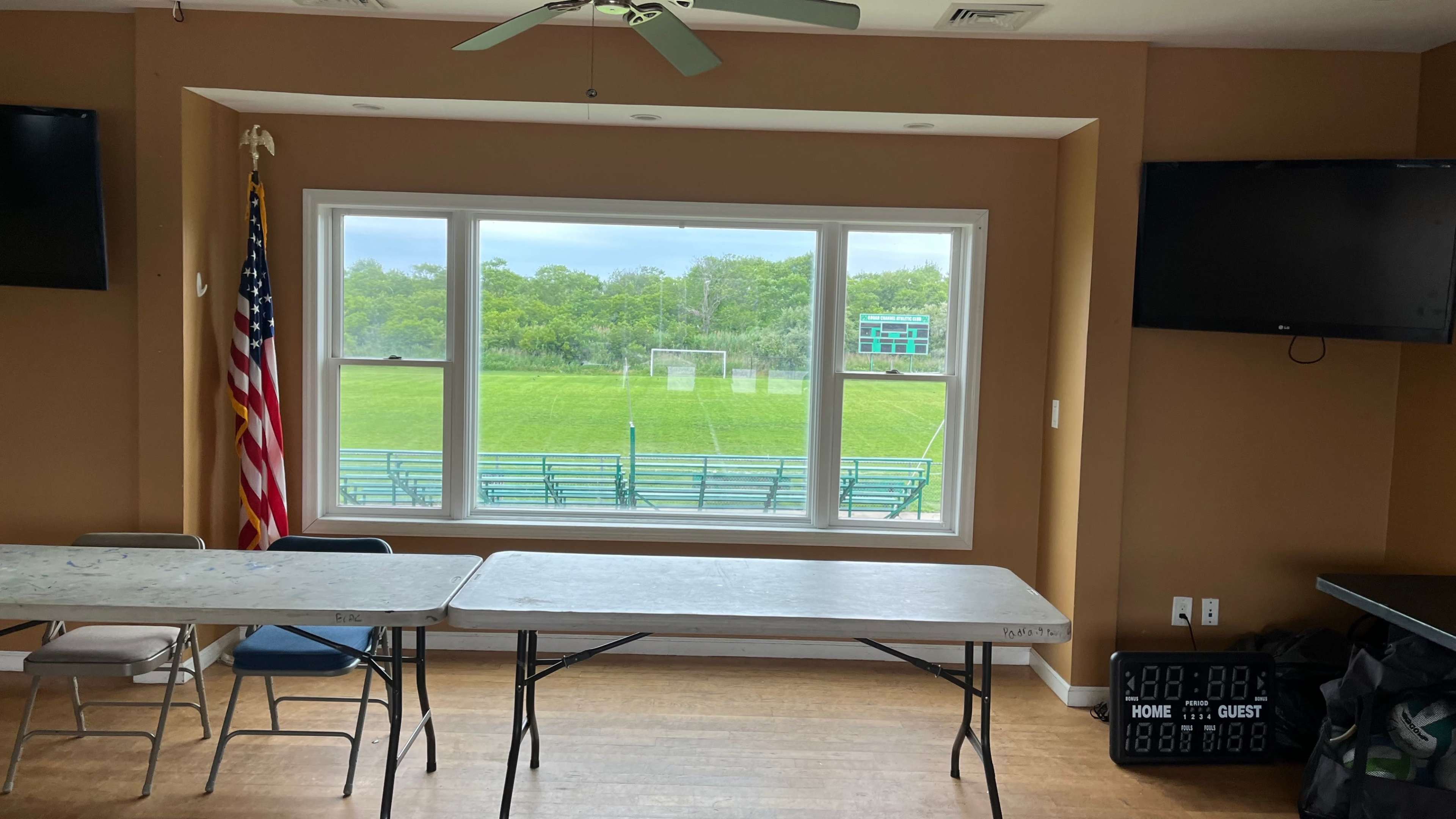 The image shows a room with two tables, an American flag, and large windows overlooking a green sports field with bleachers and a goalpost.