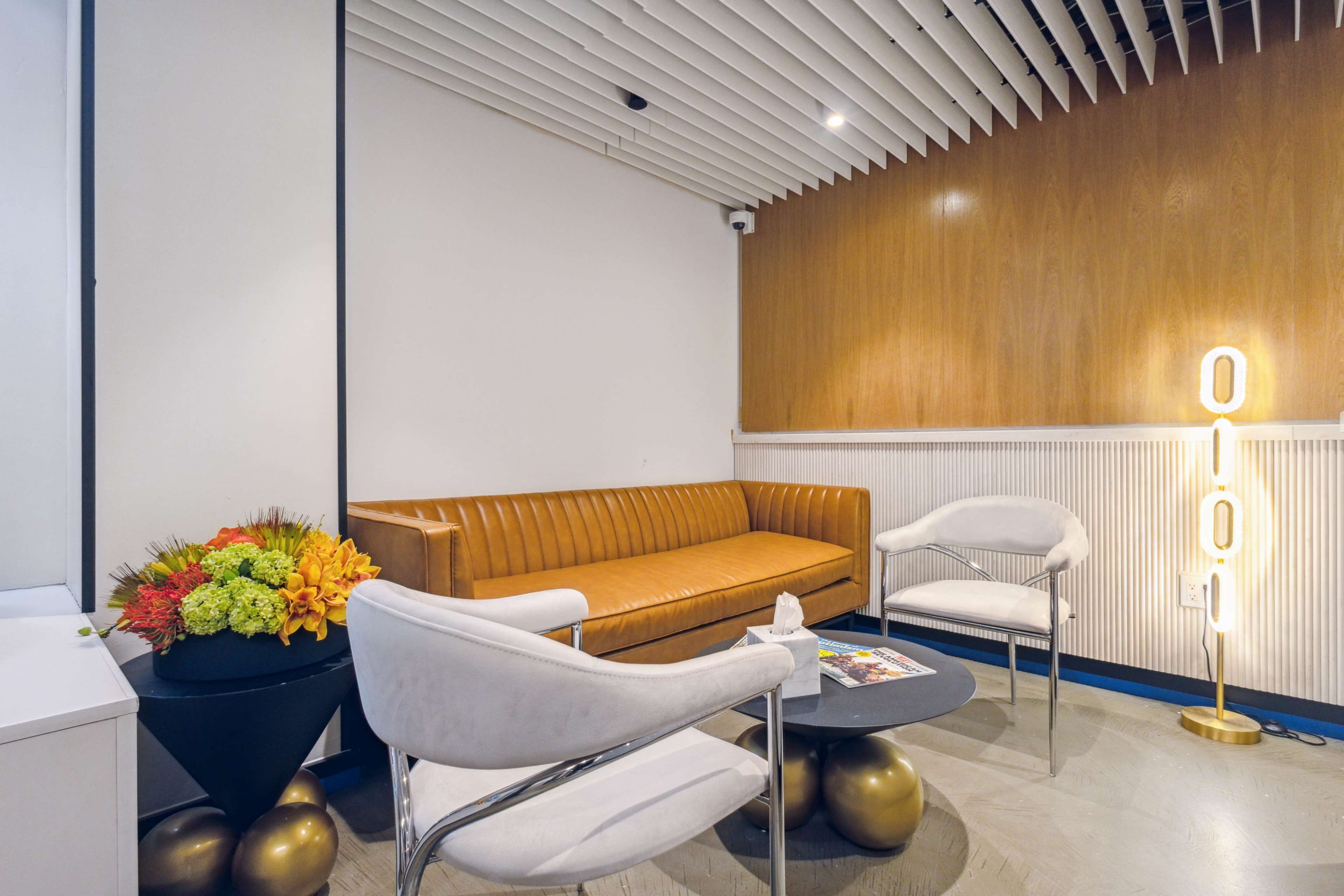 A modern waiting area with a brown leather sofa, two white chairs, a round black table, a decorative flower arrangement, and a lit floor lamp with the word "OUI" in bright letters.
