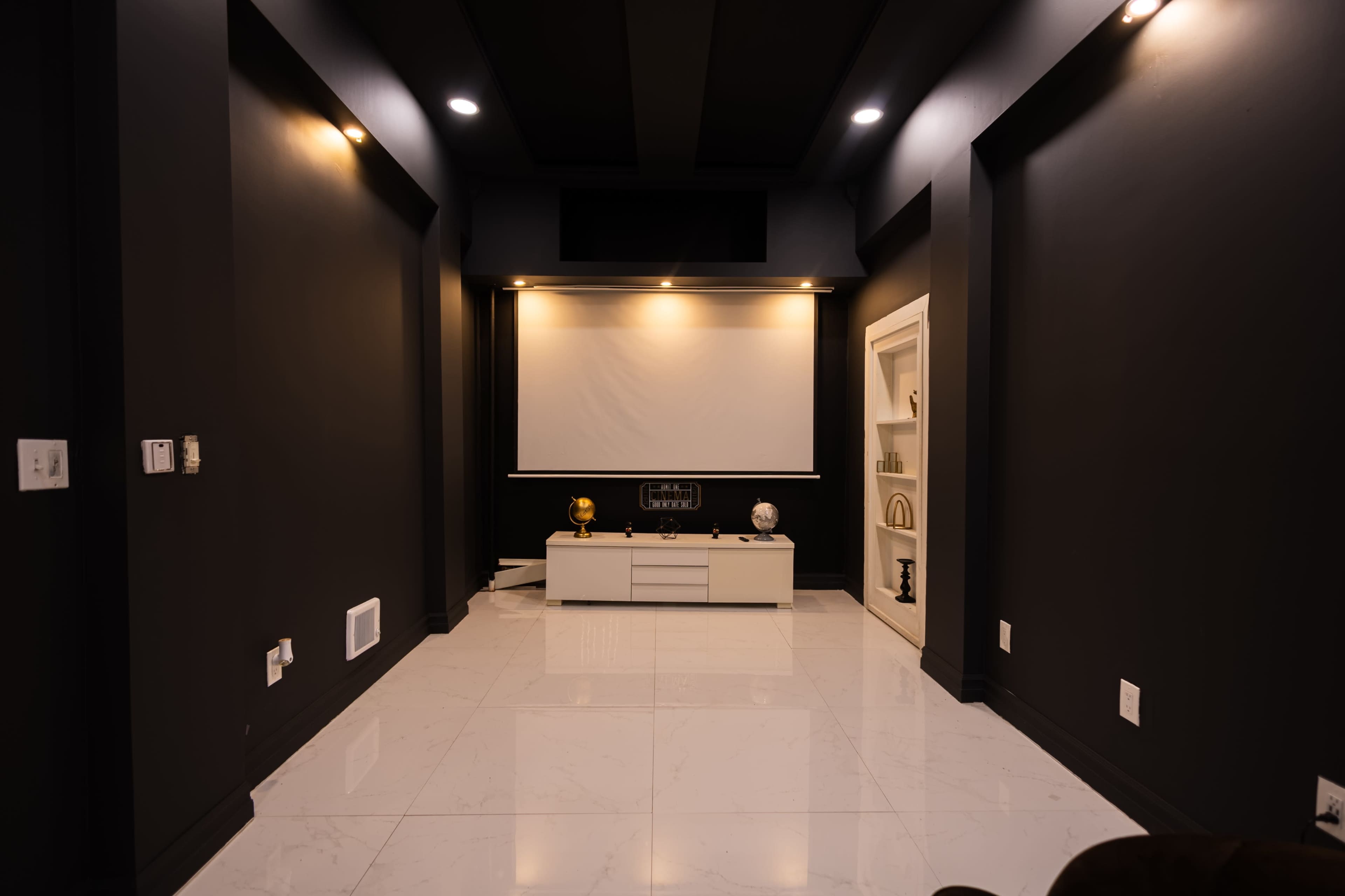 The image shows a dimly lit home theater room with black walls, a large white screen, and a minimalist white entertainment console.