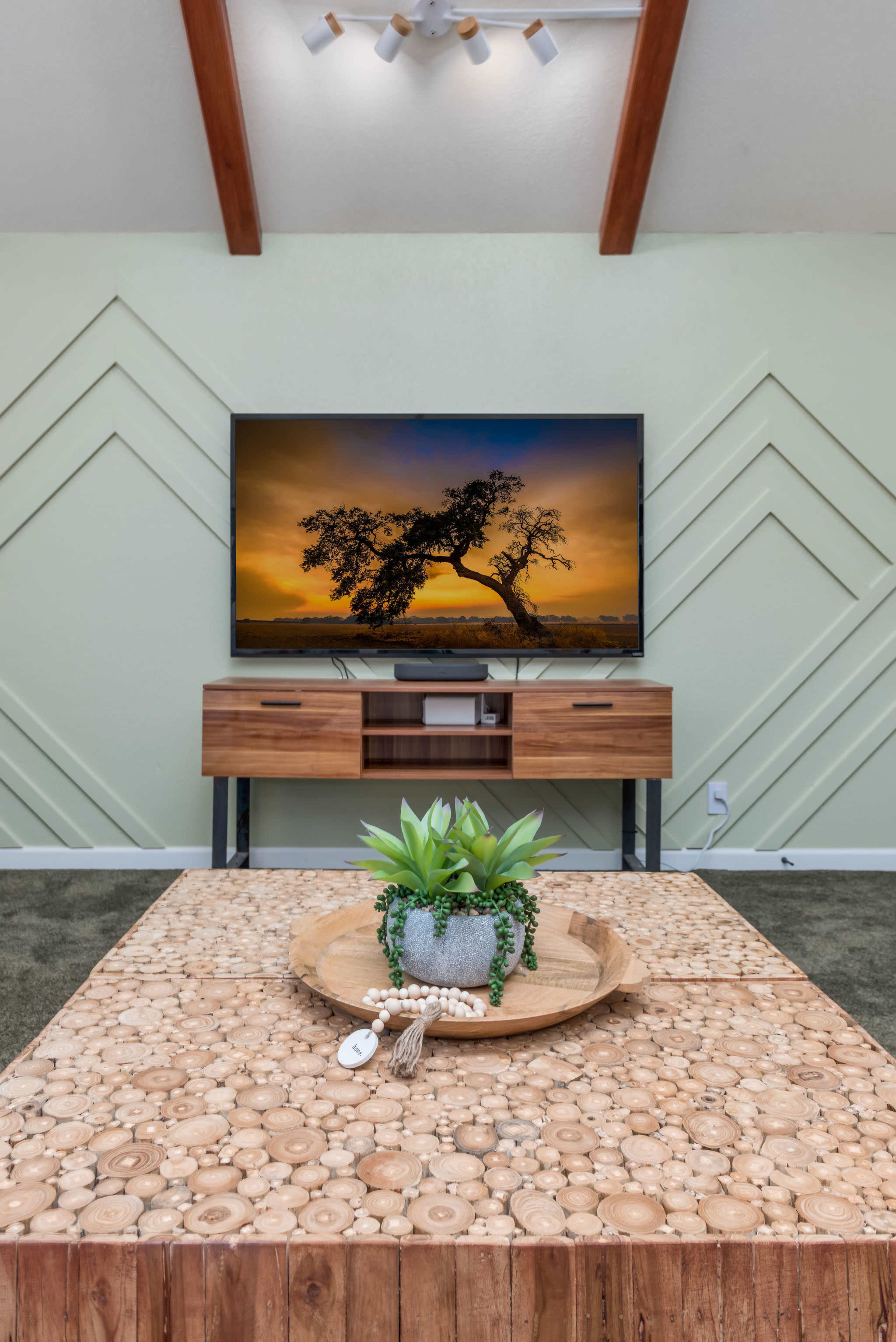 A modern living room features a wooden coffee table with a round tray and decorative plant, a media console beneath a television displaying a tree silhouette, and geometric wall accents.