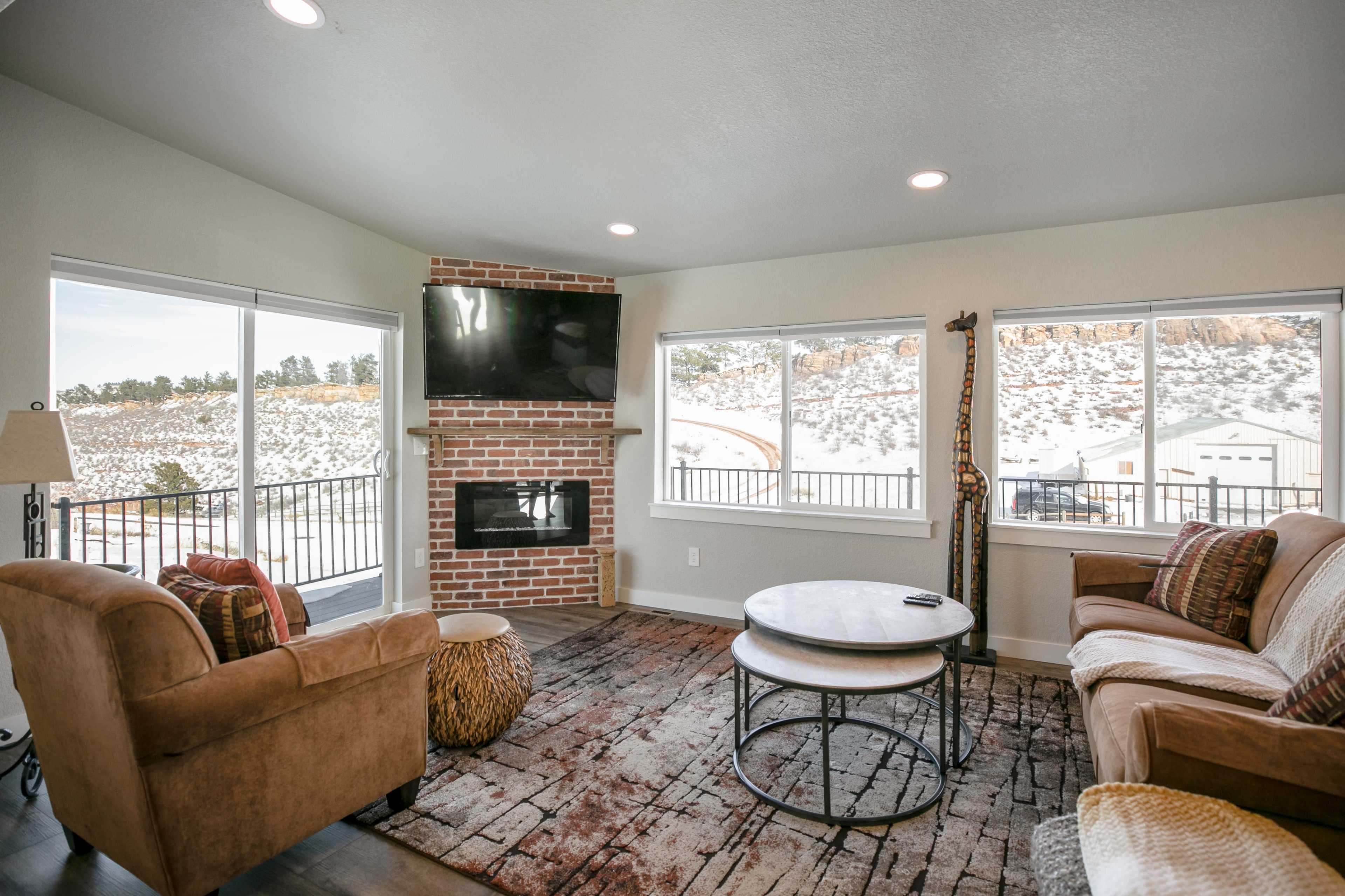 The image shows a cozy living room with a brick fireplace, two armchairs, a round coffee table, and large windows offering a view of snowy hills.