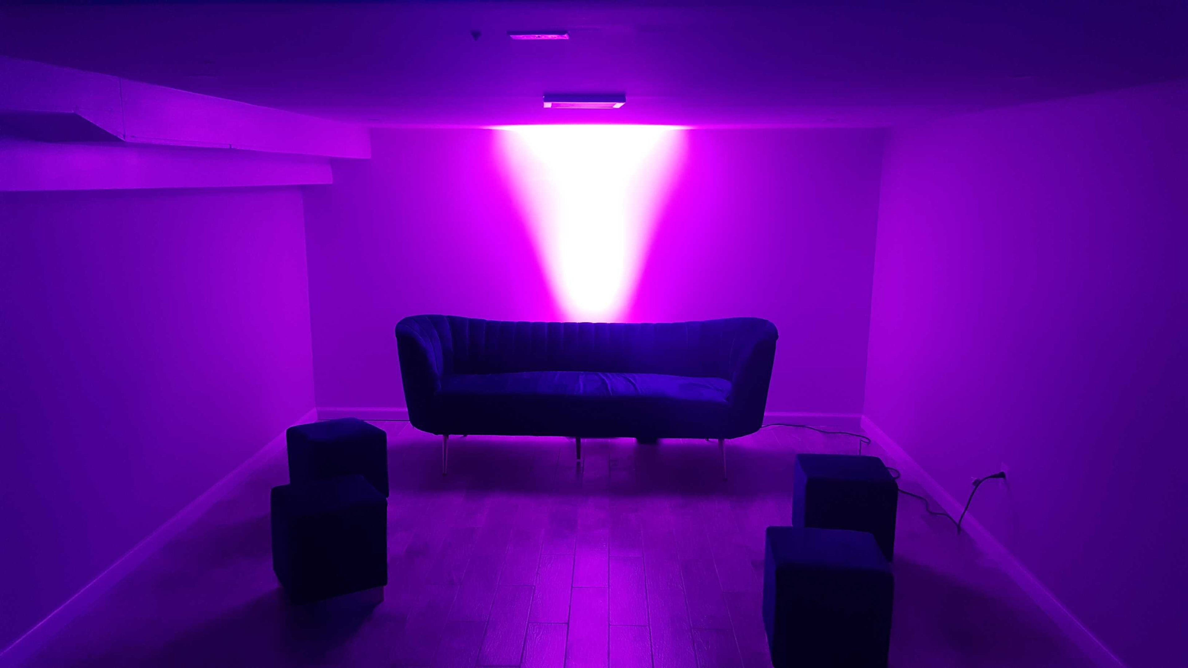 A purple-lit room features a blue couch and several small stools arranged on a wooden floor.