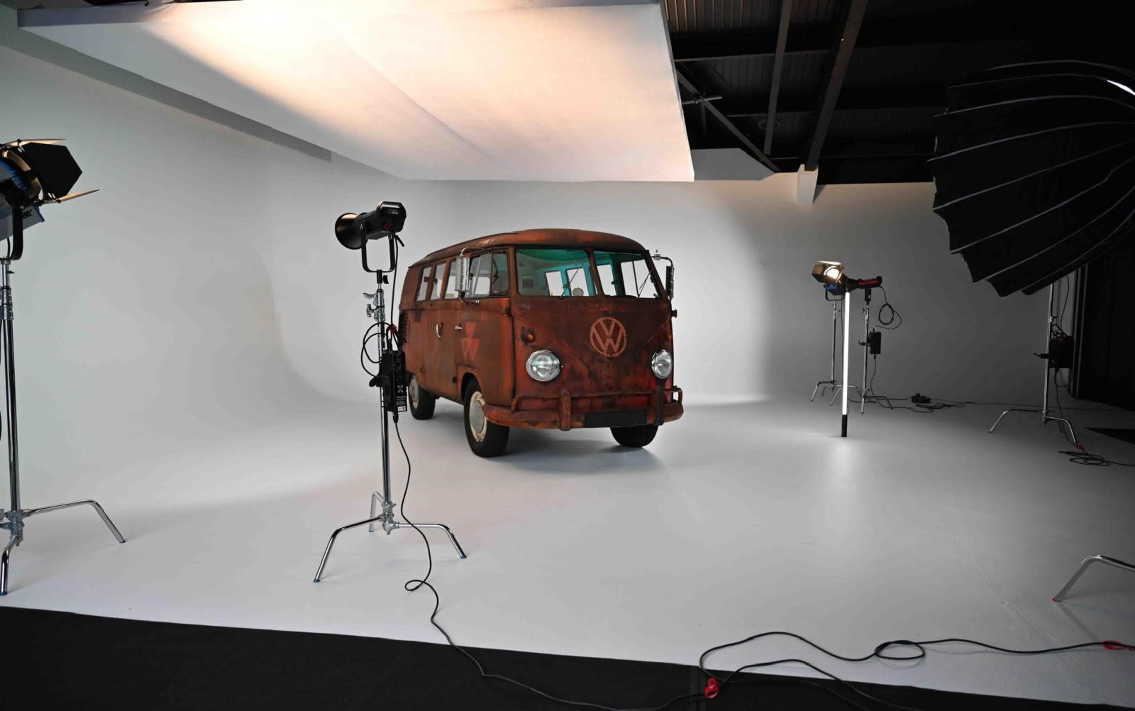 A vintage, rusty Volkswagen van is positioned in a photography studio, surrounded by studio lighting equipment on a plain backdrop.