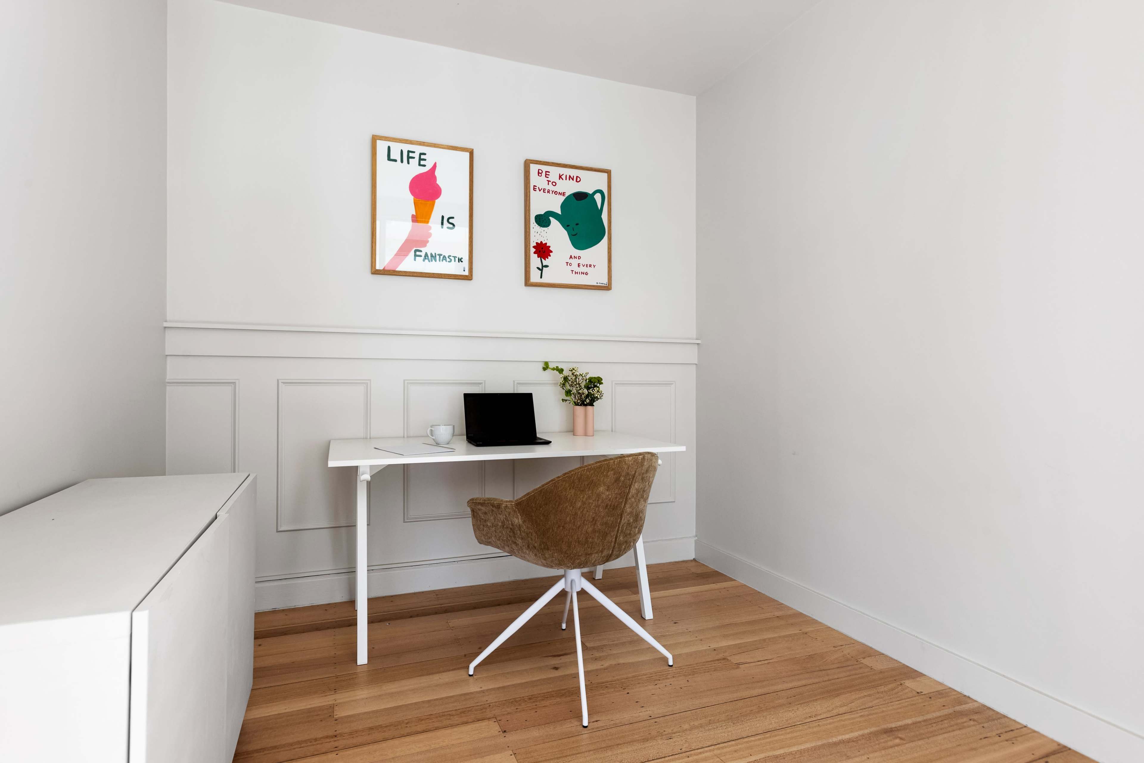 The image shows a minimalist home office space with a white desk, a chair, and two framed posters on the wall.