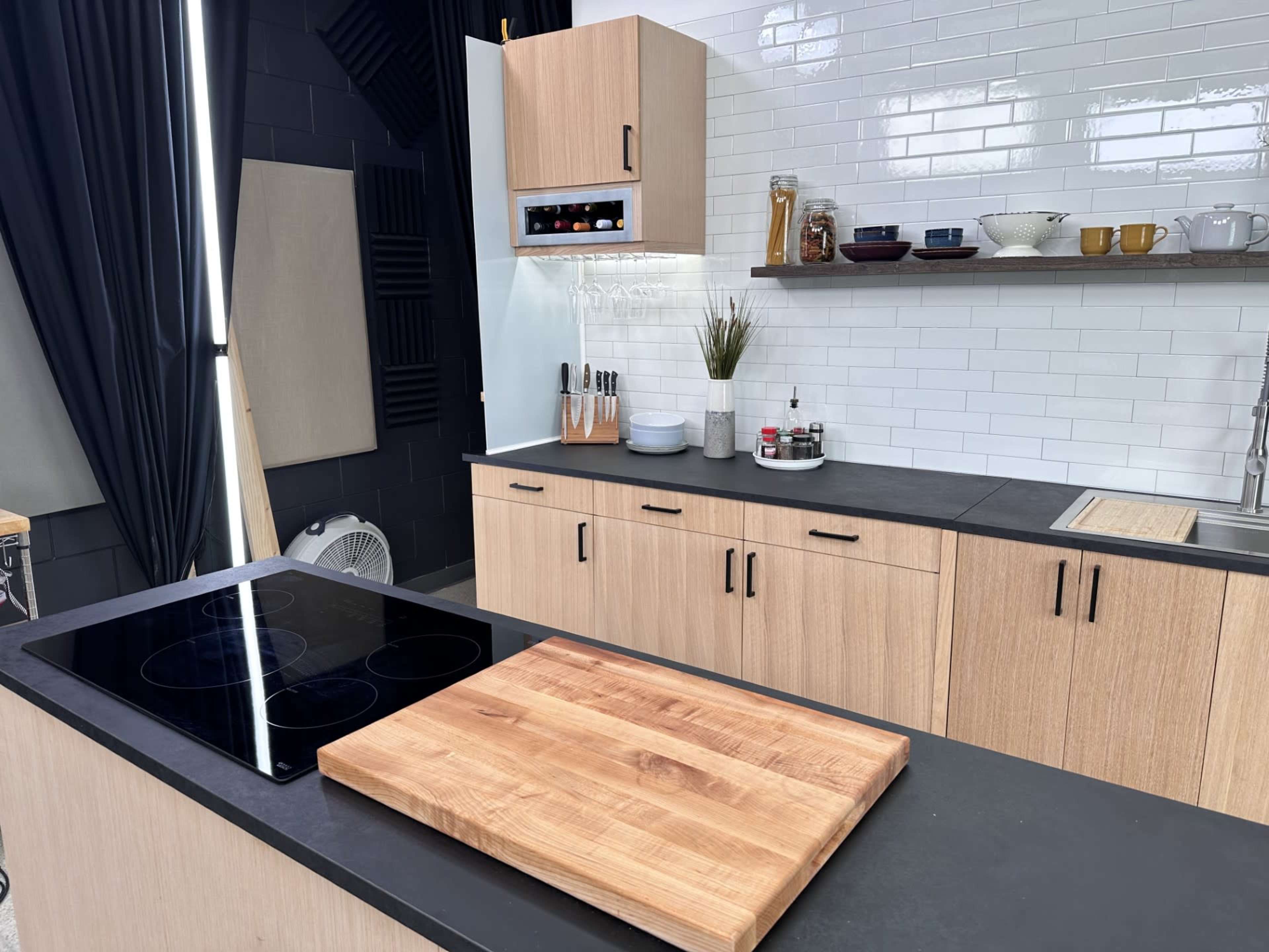 The image shows a modern kitchen with wooden cabinets, a black countertop, an induction cooktop, and a spacious cutting board.