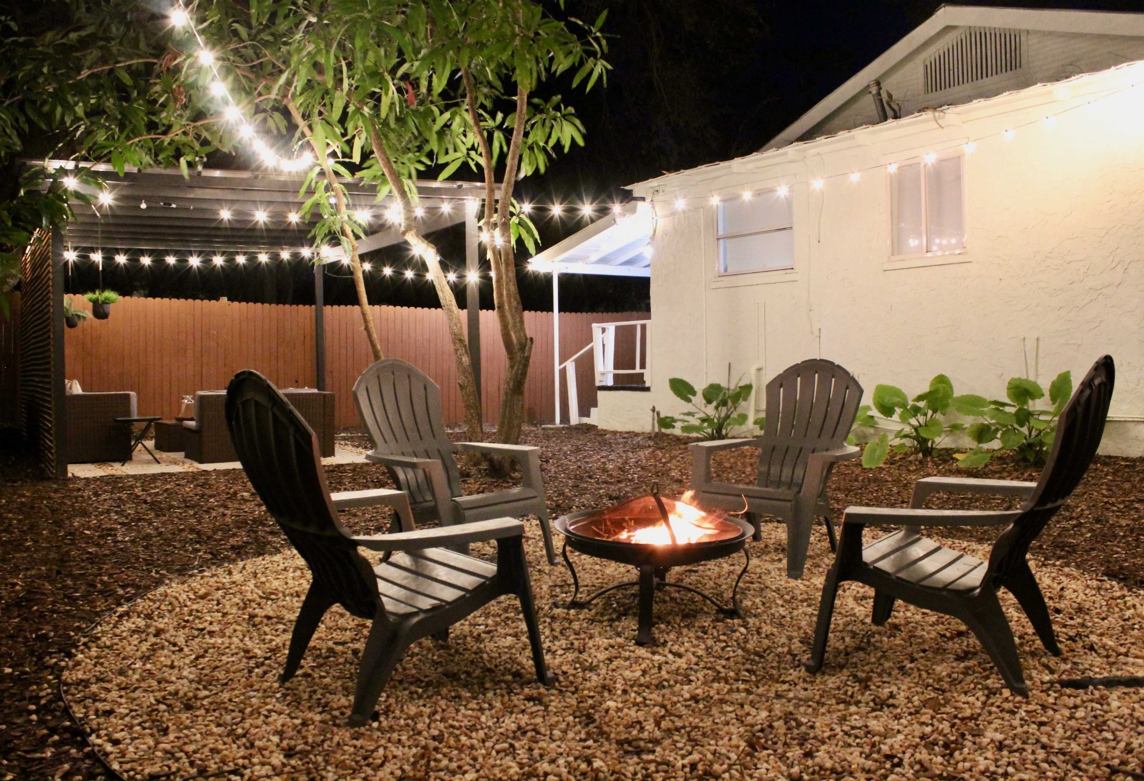 A circular arrangement of four adirondack chairs surrounds a fire pit in a backyard illuminated by string lights.