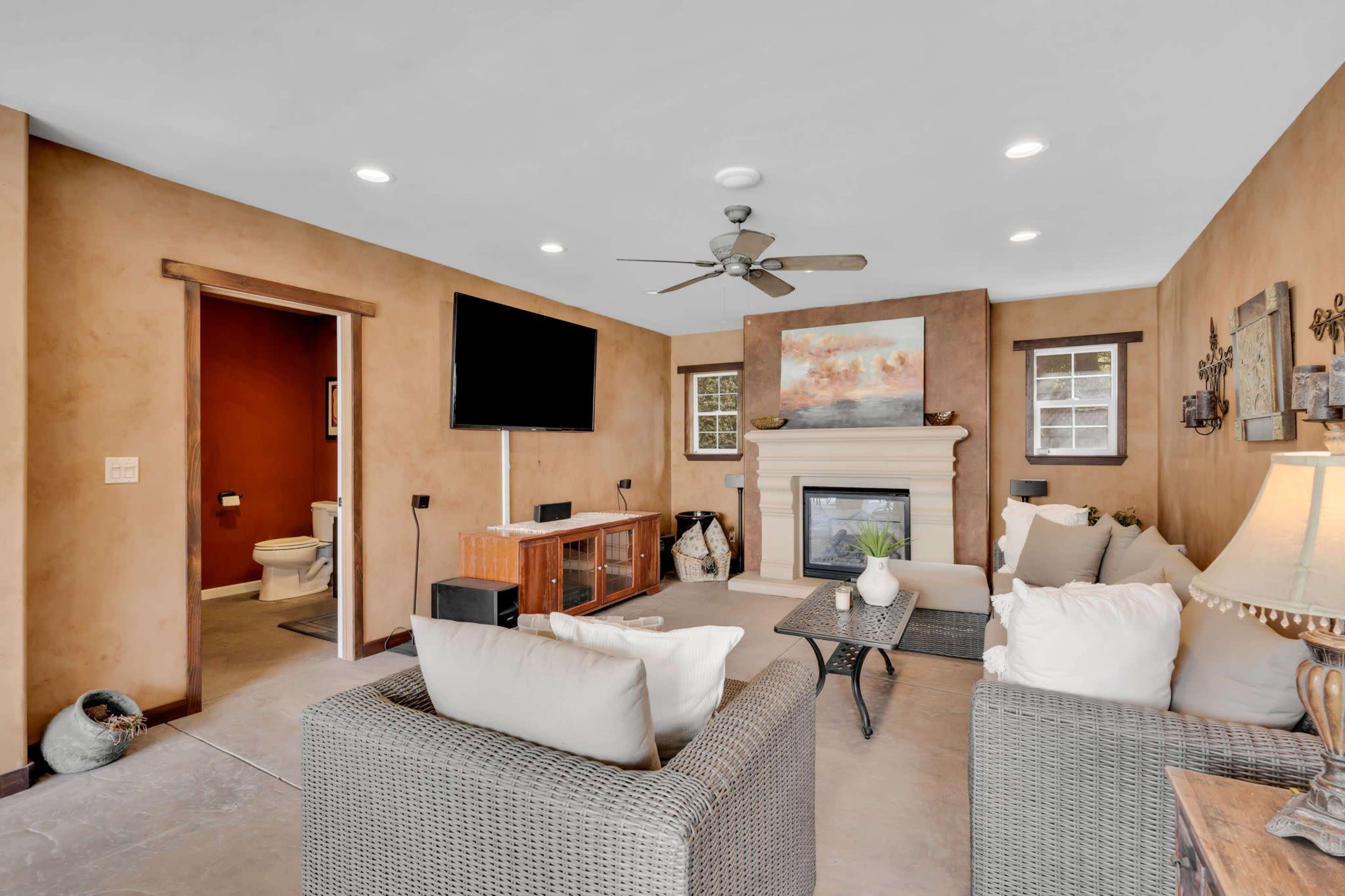 The living room features a seating area with light-colored couches, a television mounted on the wall, and a fireplace, with an adjacent room visible through a doorway.