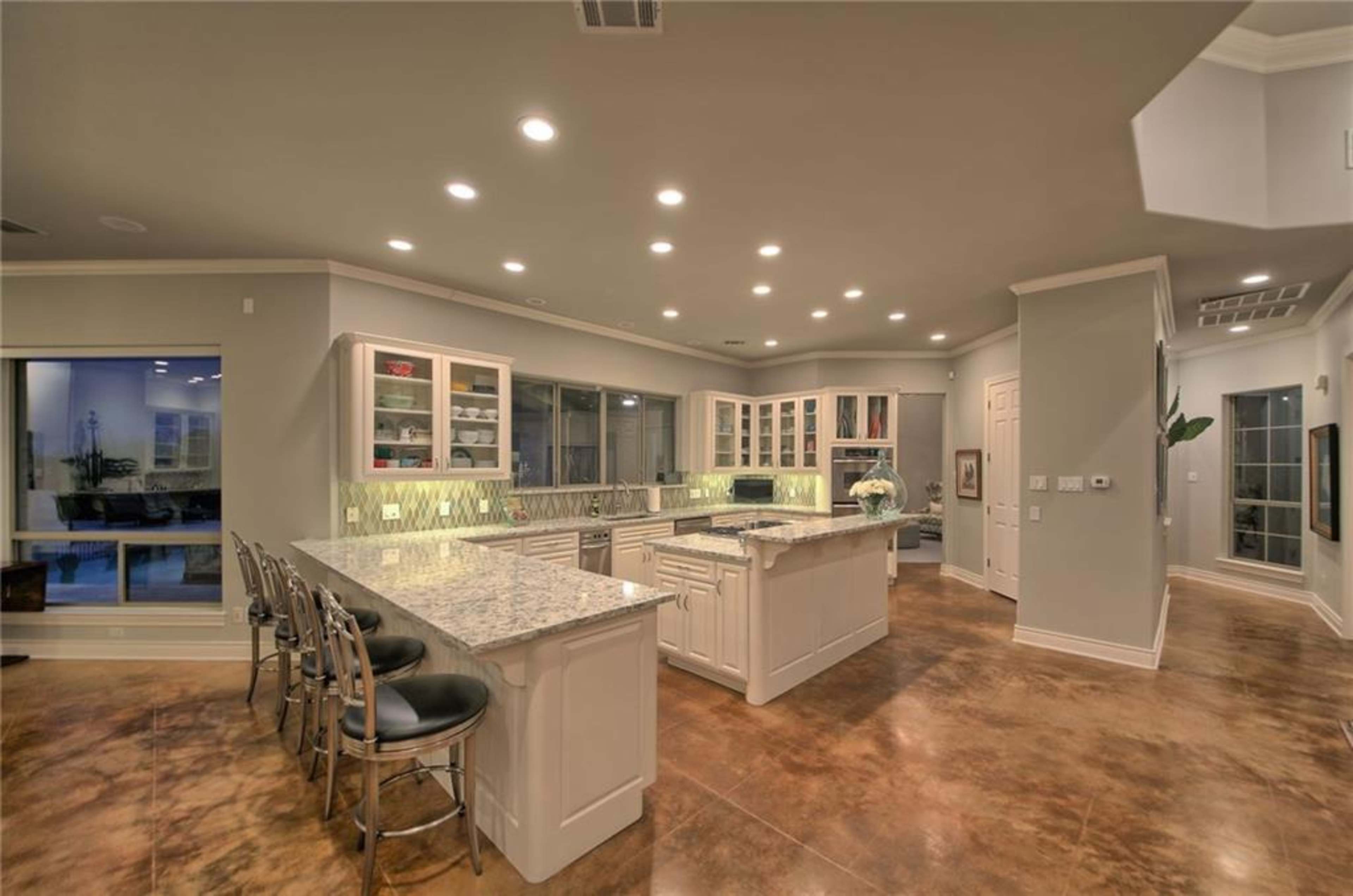 The kitchen features a large island with granite countertops, white cabinetry, and stainless steel appliances, illuminated by recessed lighting.