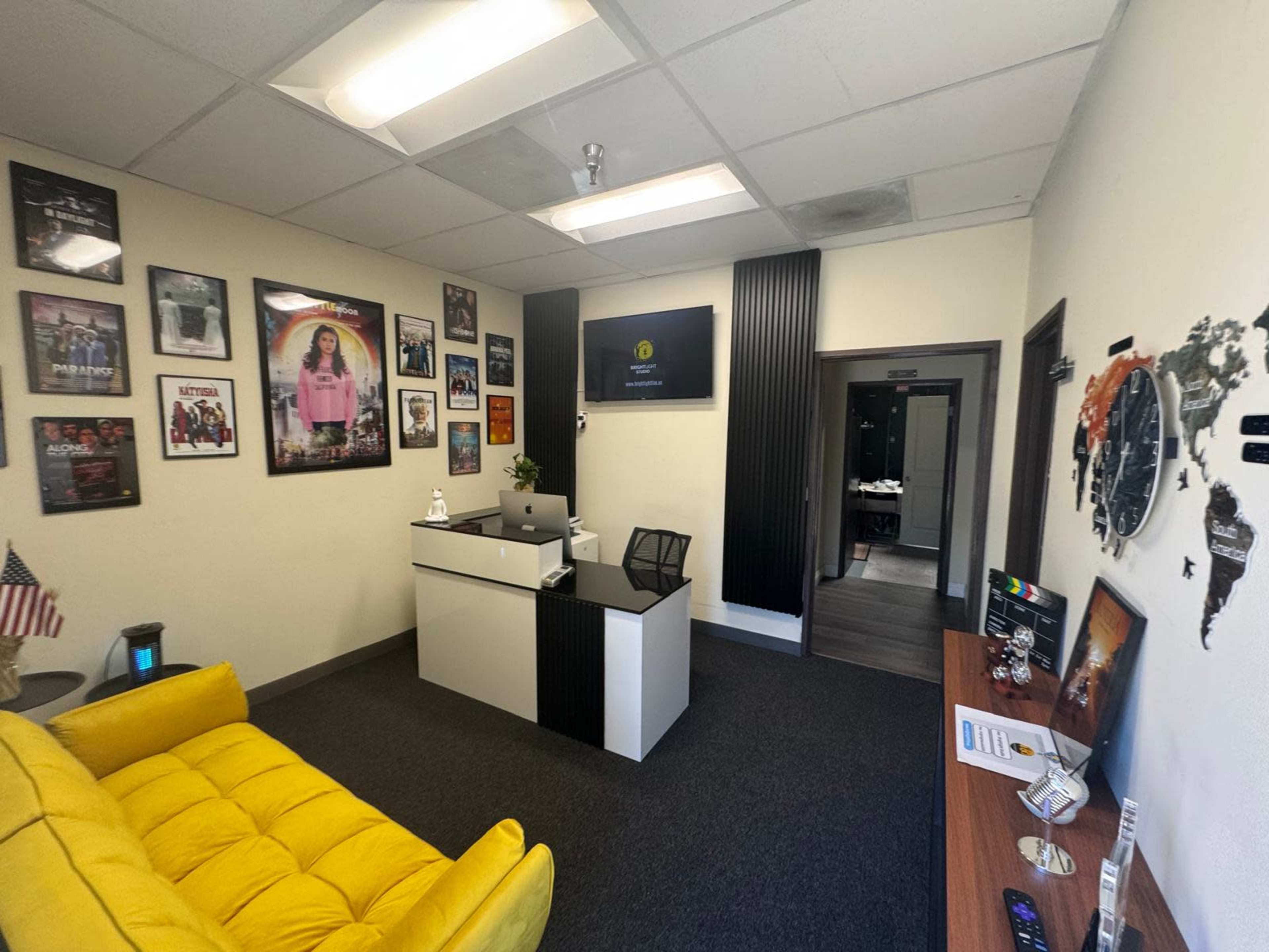 The image shows a modern office space with a yellow couch, a reception desk with a computer, and decorated walls featuring various framed photos and a wall map.