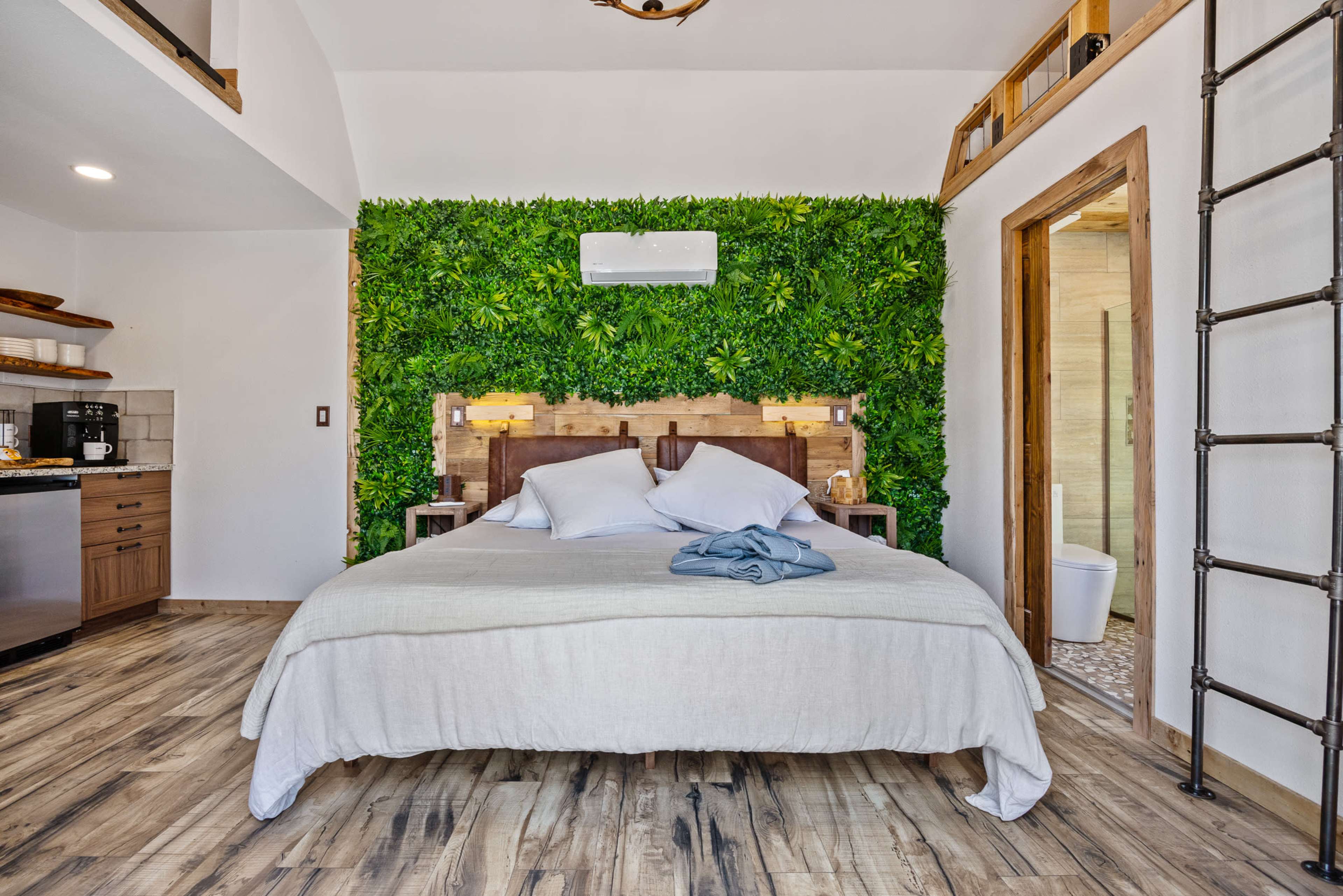 A modern bedroom features a bed with white linens and a green plant wall, alongside a kitchenette and a bathroom area.