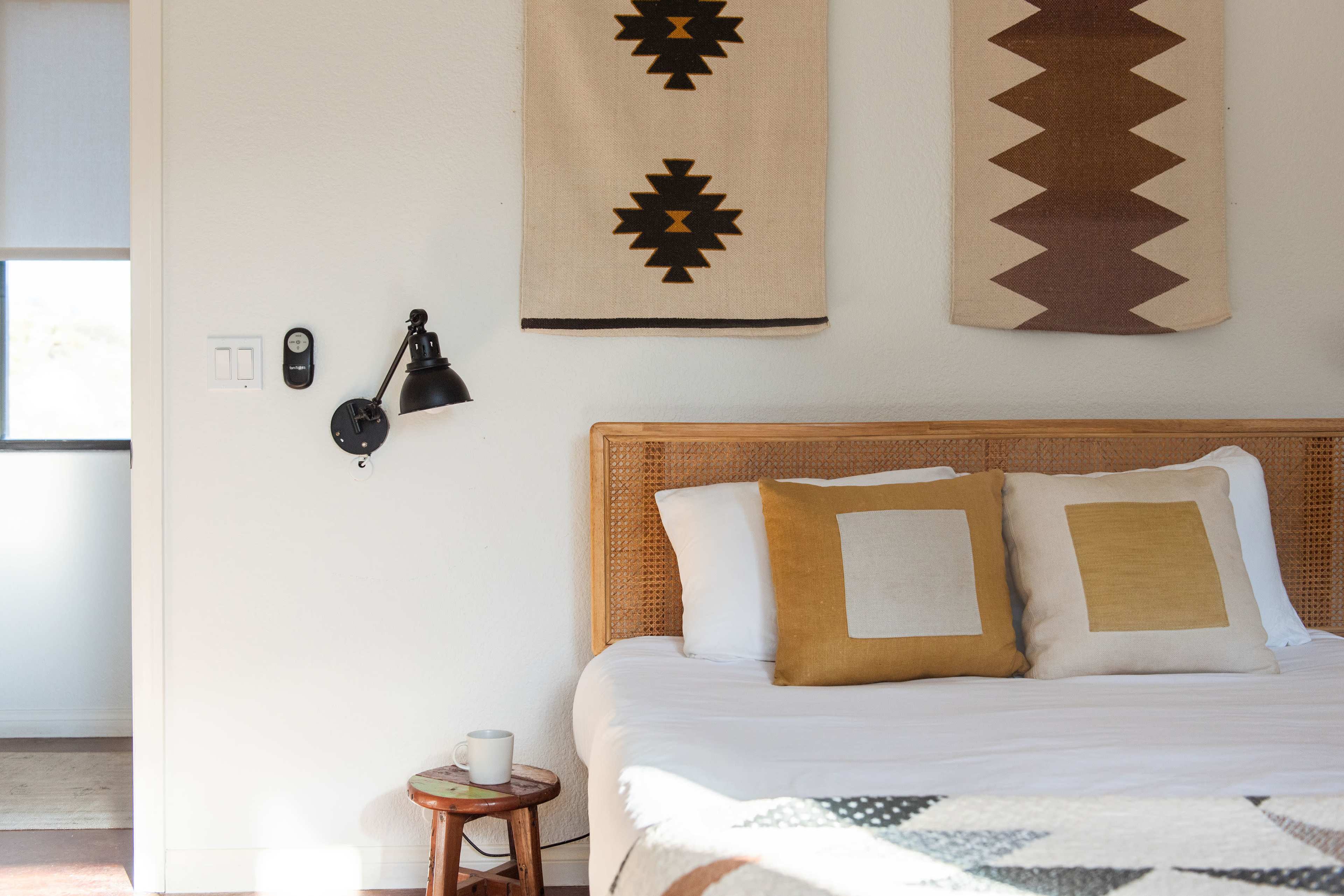 The image features a minimalist bedroom with a wooden bed, two decorative pillows, and wall hangings above the bed, alongside a small side table with a cup.