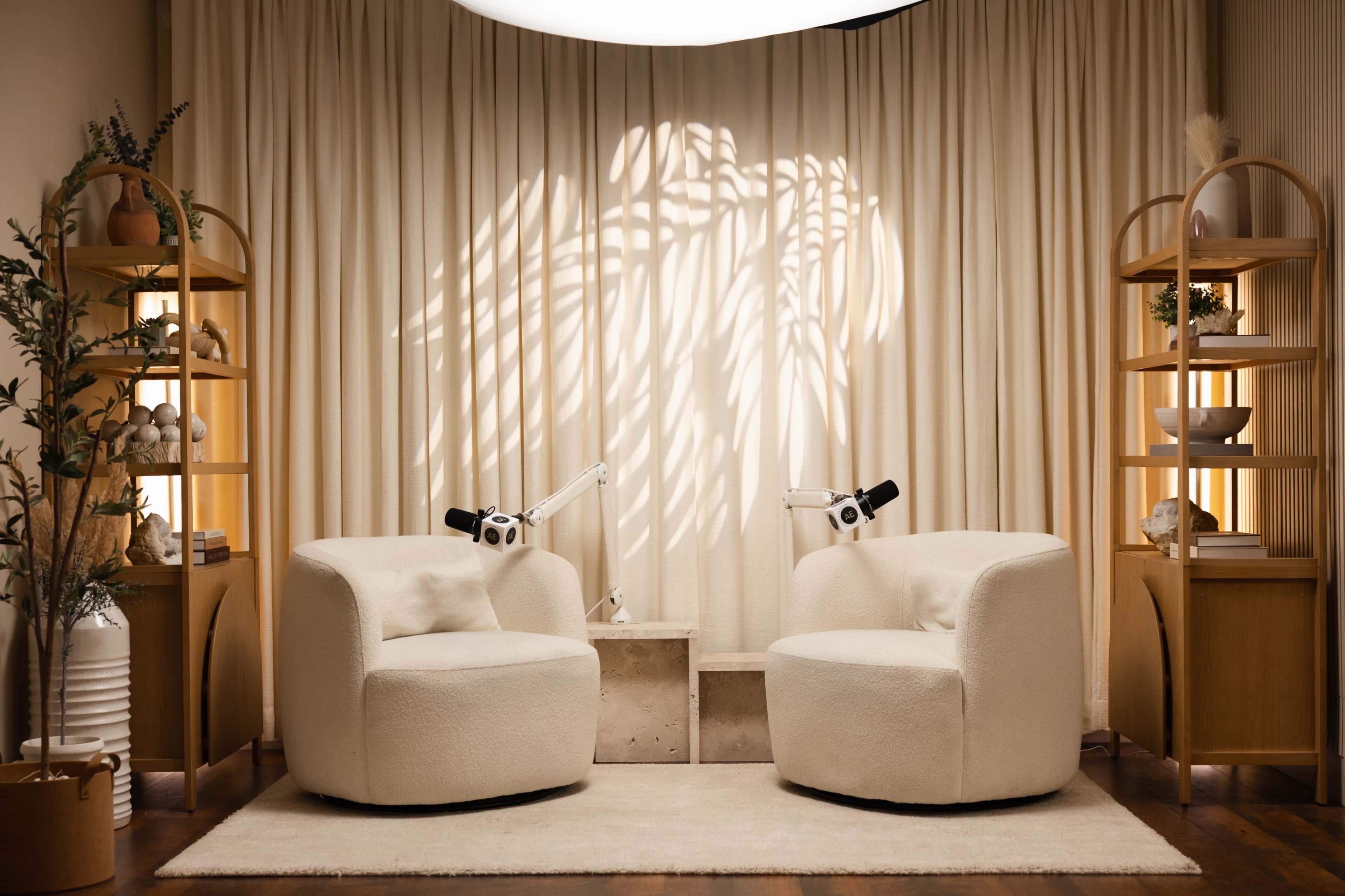 The image features a cozy, well-lit seating area with two round, plush chairs facing each other, flanked by wooden shelves and accented by soft textile curtains.
