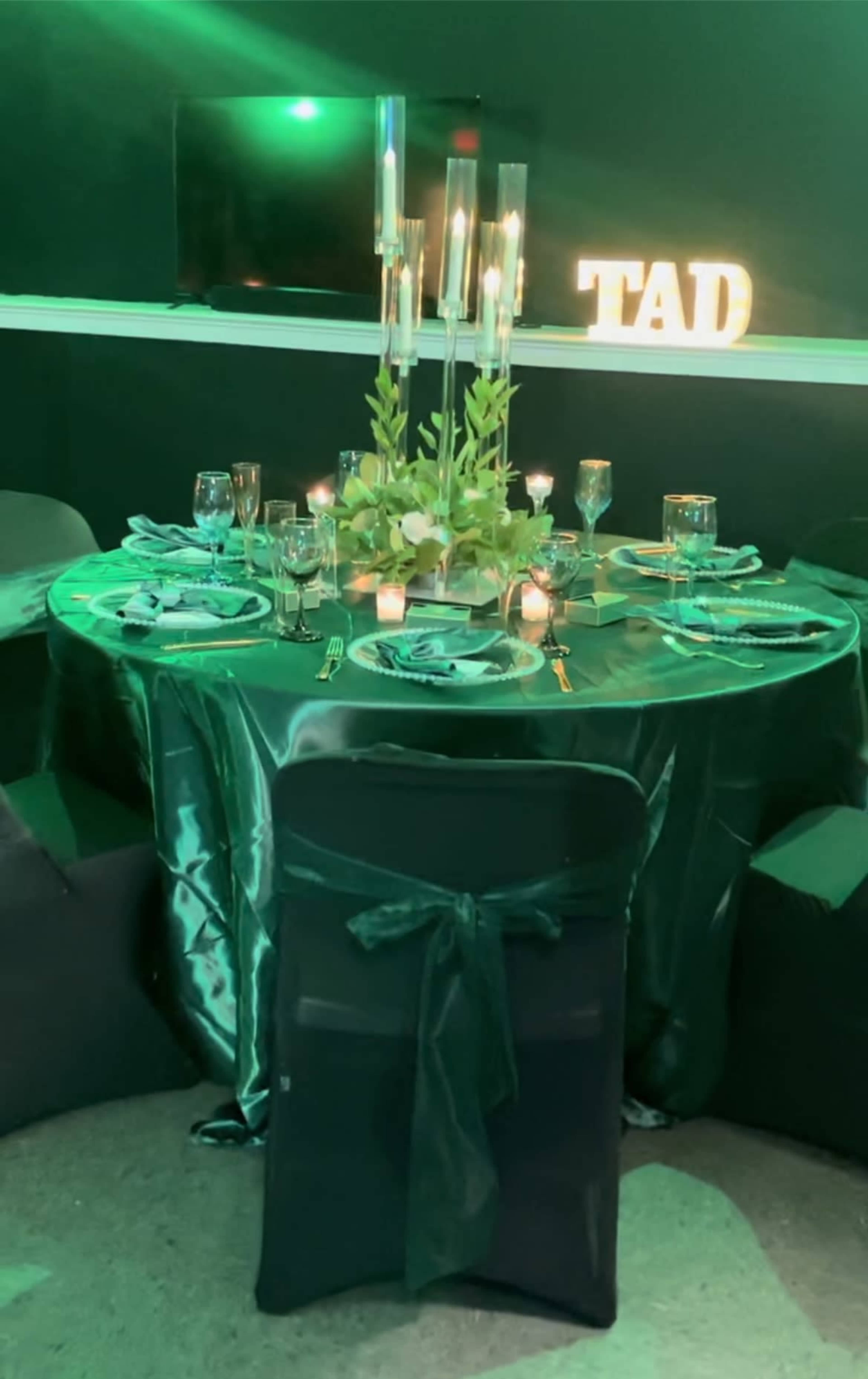 A round table set for an event, featuring a green tablecloth, elegant place settings, candles, and a decorative arrangement of plants, with illuminated letters spelling "TAD" in the background.