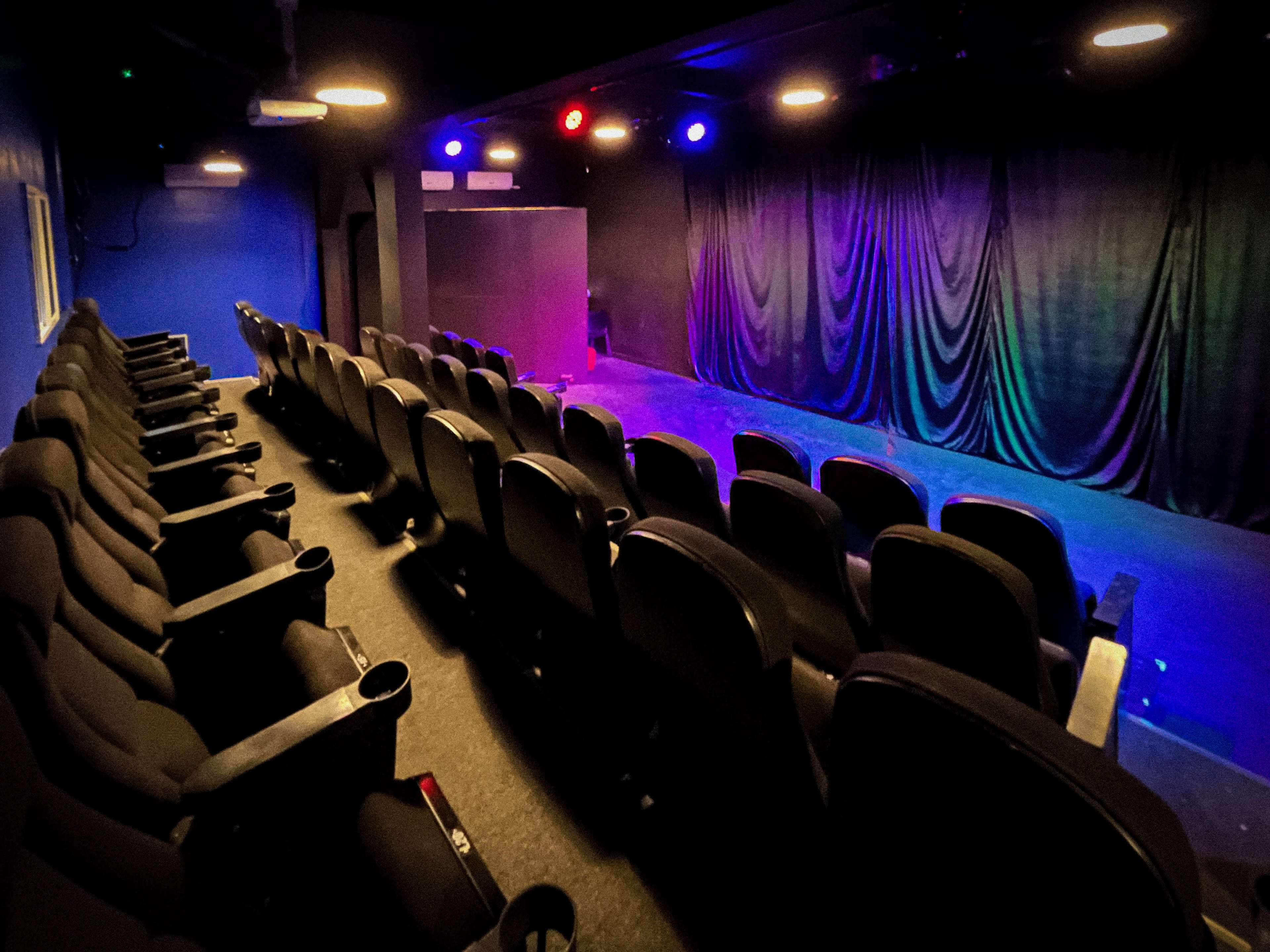 The image shows an empty theater with rows of black seats facing a stage illuminated by colorful lights.
